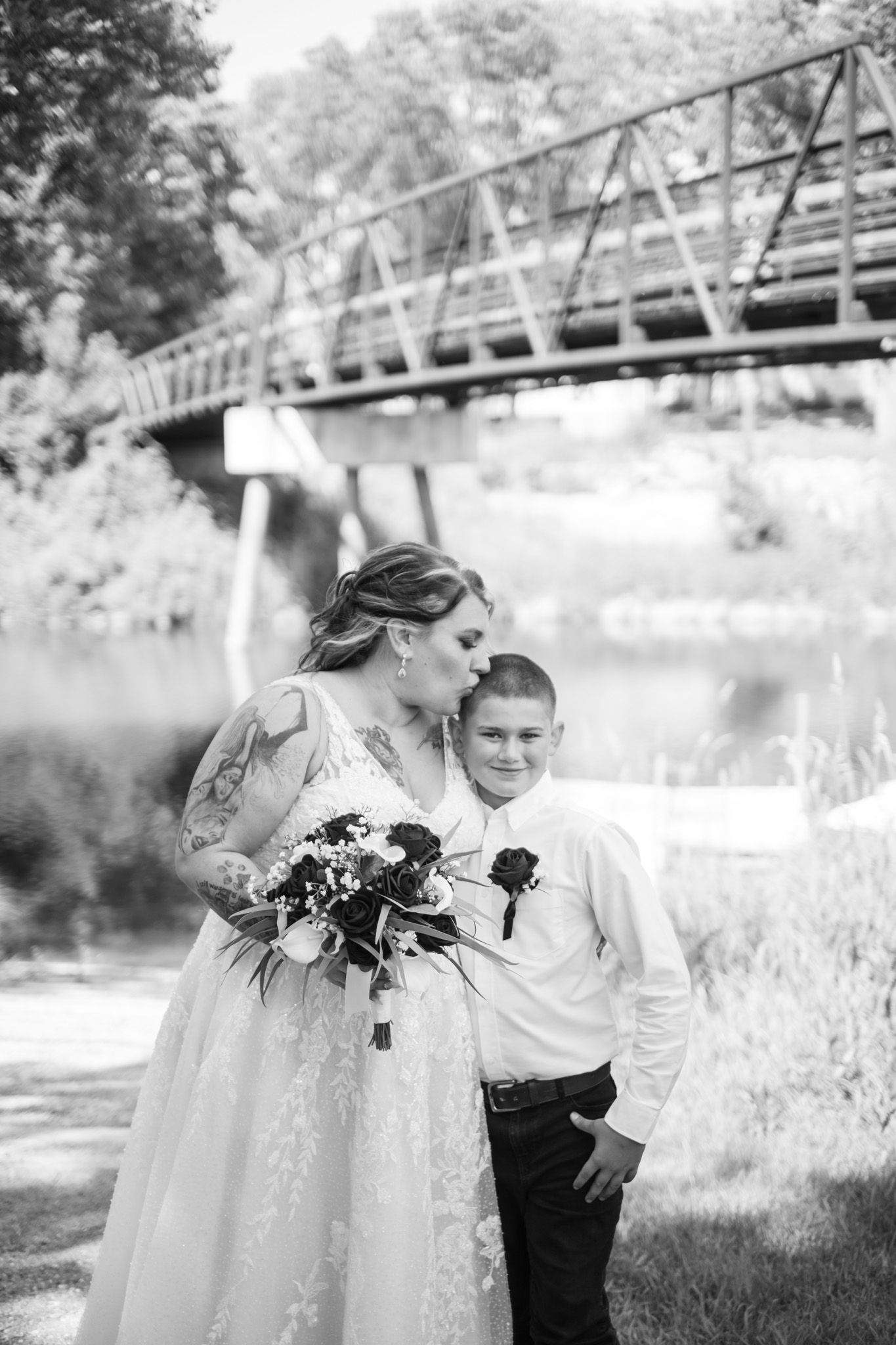 Black and white wedding photo near a rustic bridge over water showing a tender moment between two people.