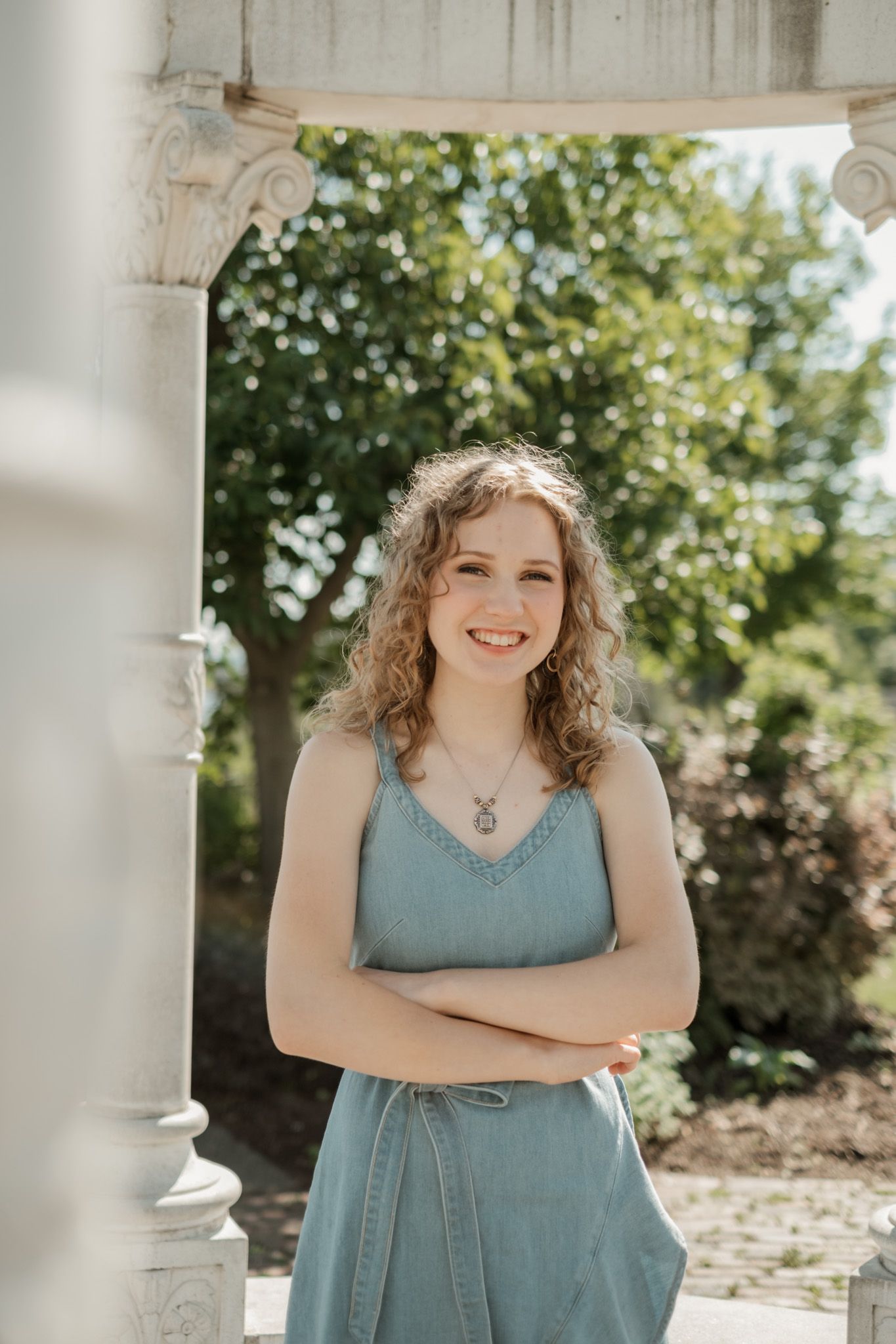 Young woman in blue denim dress smiles while standing on sunny porch against green foliage background.