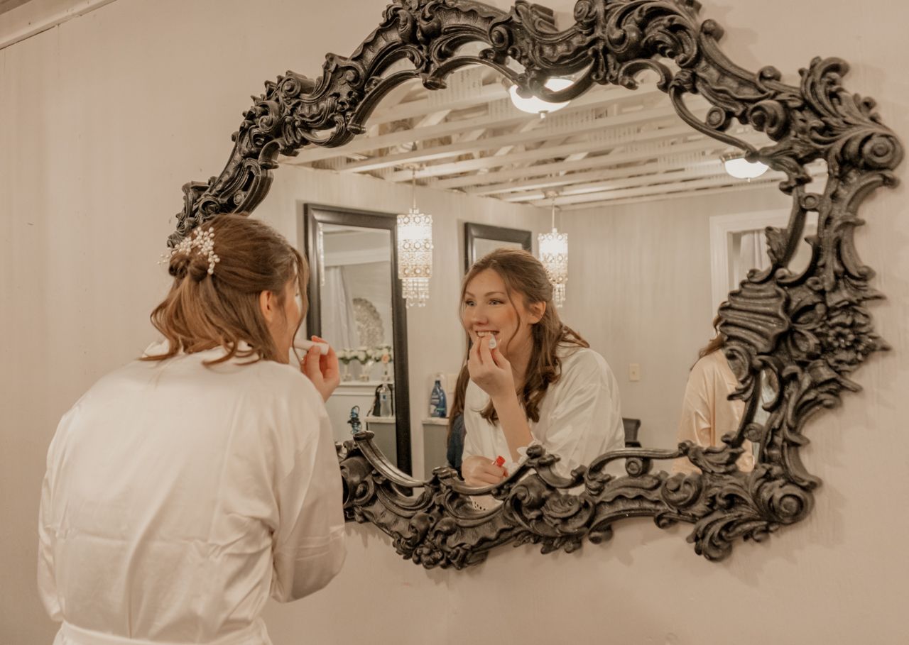 A person in a white robe is reflected in an ornate vintage mirror while getting ready.