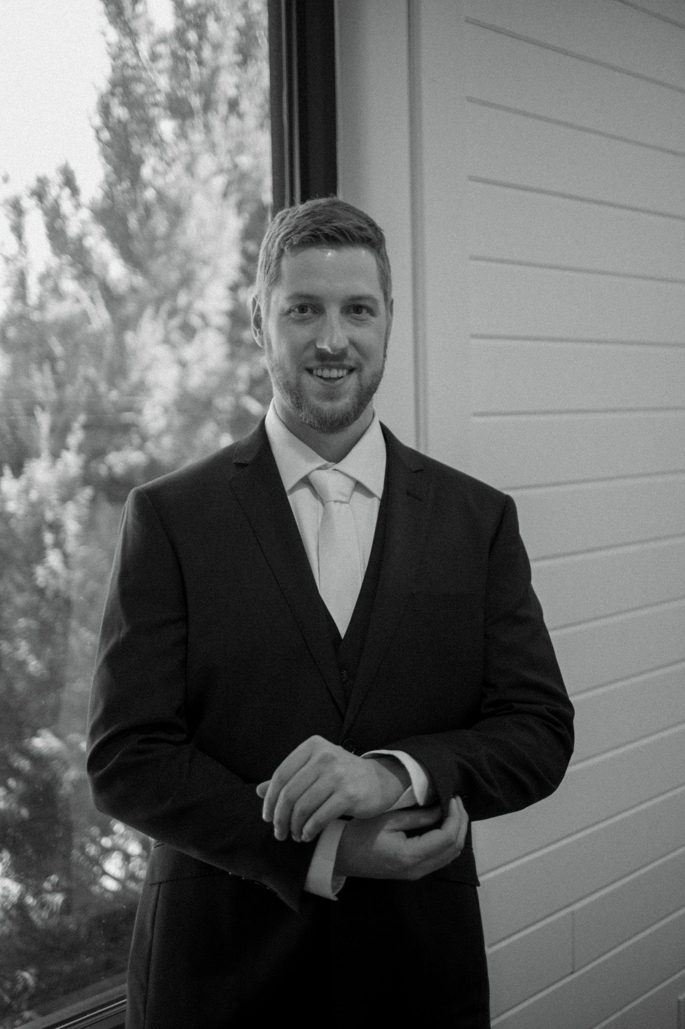 A formally dressed man in a black suit and white tie smiles while standing against a light wall.