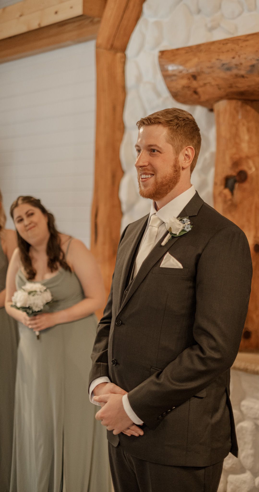 A smiling groom stands at the altar wearing a dark suit and white boutonniere with a bridesmaid behind him.