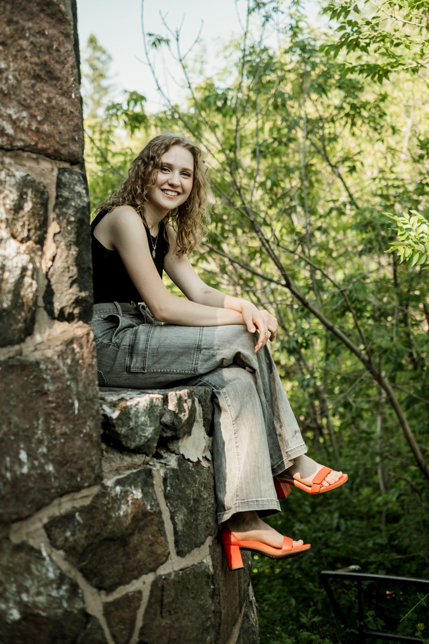 Someone sits on a rock formation wearing jeans and bright orange shoes in a lush green forest setting.