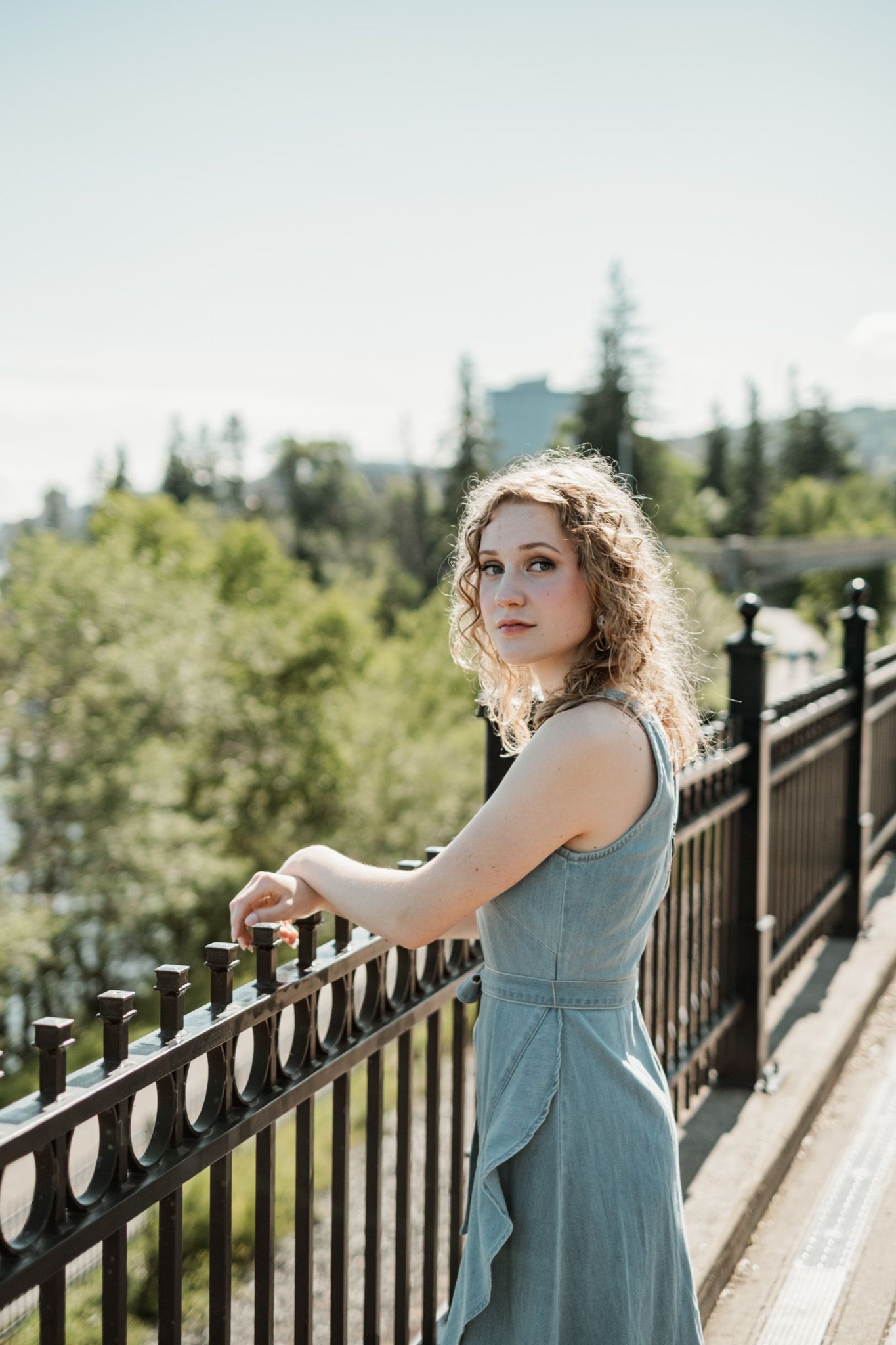 Person in a flowing seafoam dress poses on bridge with trees and buildings in background during sunny day.