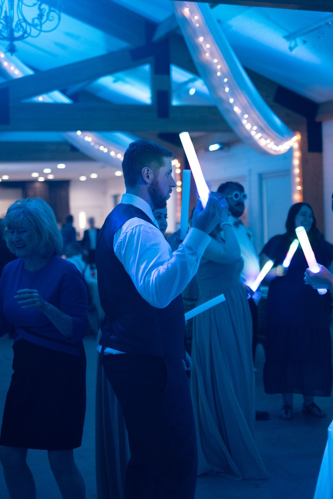Wedding reception guests holding light sticks under blue lighting and string-lit ceiling beams.