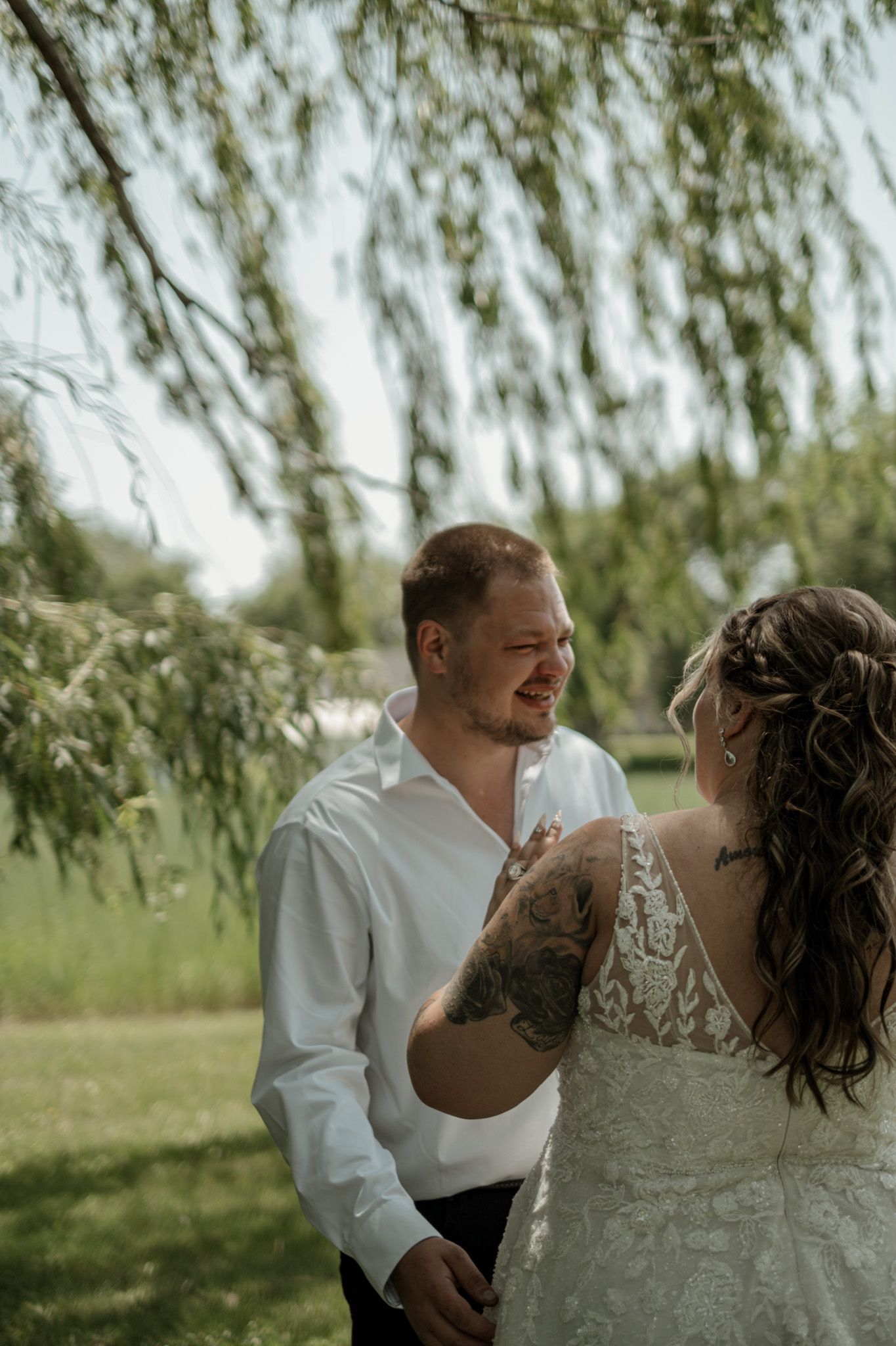 A romantic wedding couple shares joyful laughter together under a willow tree in a natural outdoor setting.