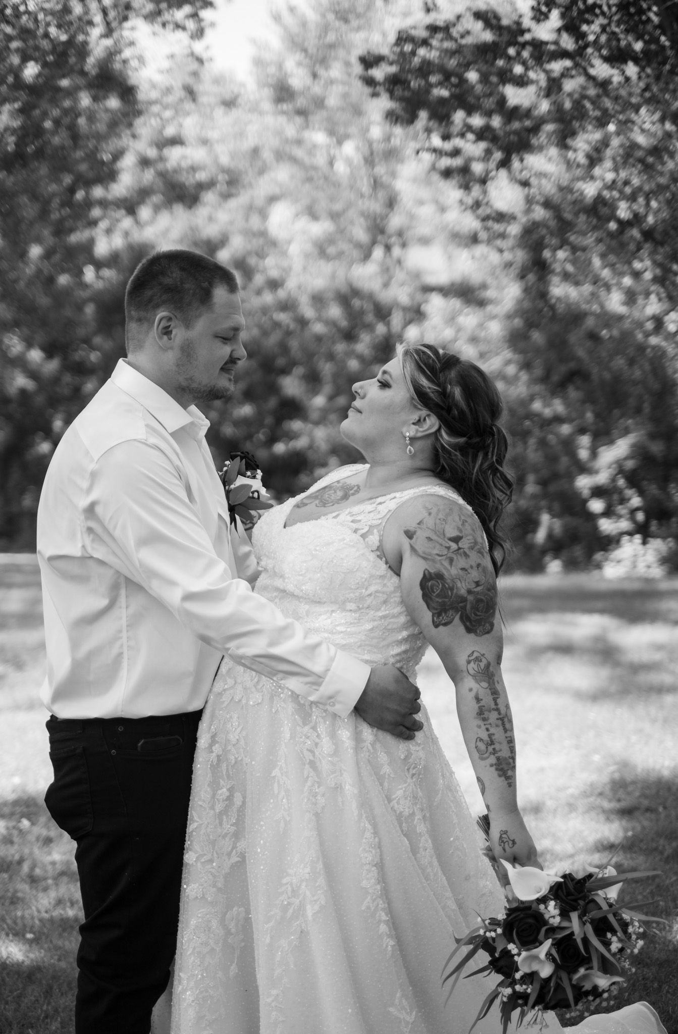 Black and white wedding portrait of a couple in formal attire embracing outdoors under trees.