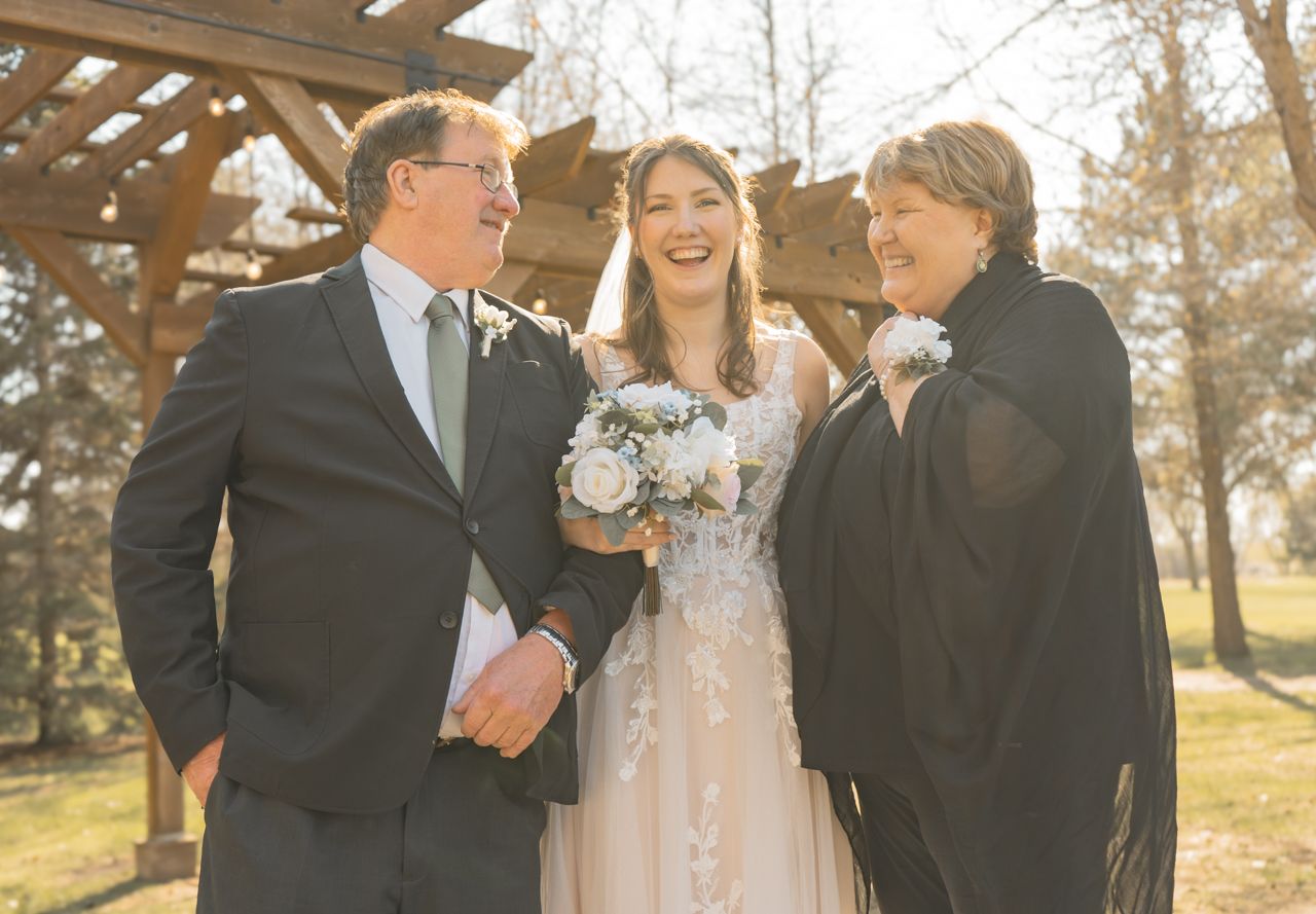 Wedding party sharing a joyful moment together under a wooden pergola in soft sunlight.