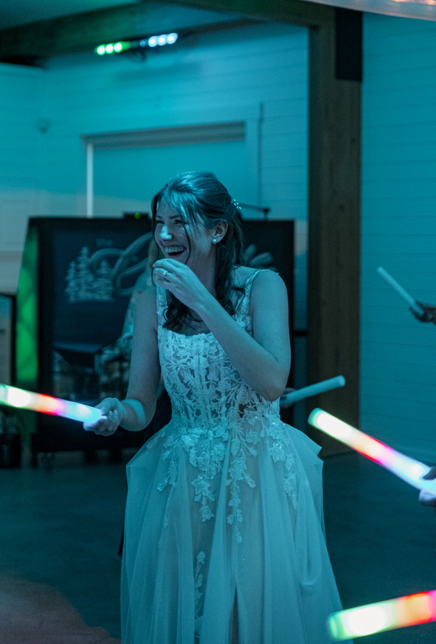 A person in a white lace dress smiles while holding glowing light wands in a darkened room with teal lighting.