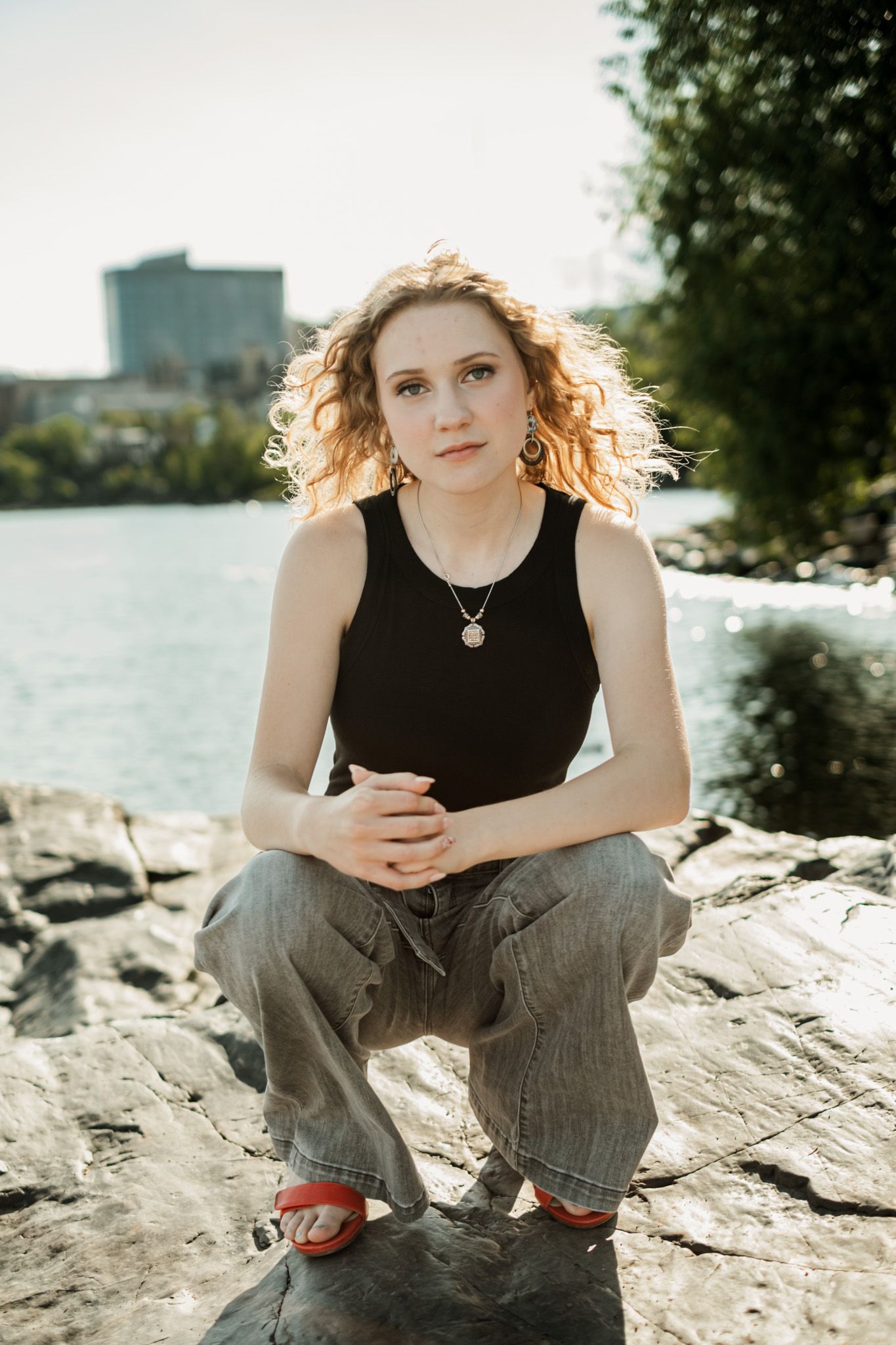 Portrait by the water with sunlit curly hair wearing black tank top and cargo pants sitting on rocks.