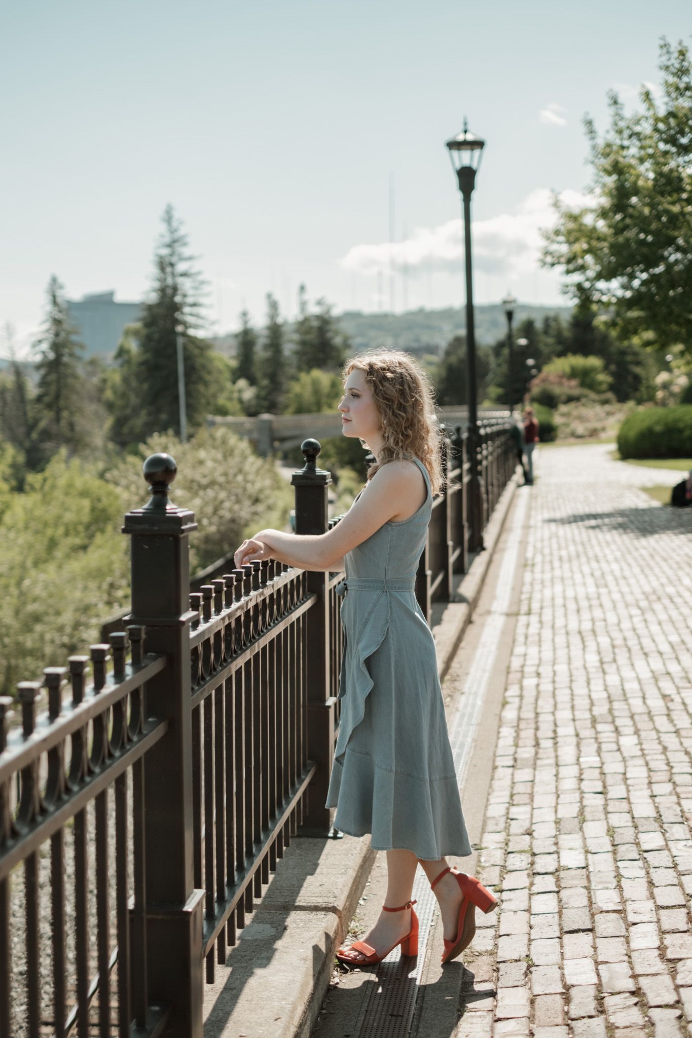 A stylish figure in a teal dress poses elegantly along a brick pathway with black iron railings on a sunny day.
