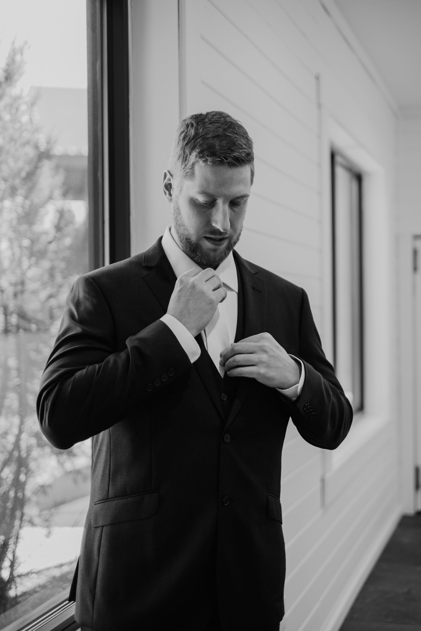 Man in dark suit adjusts his tie while getting ready by a window in black and white photography.