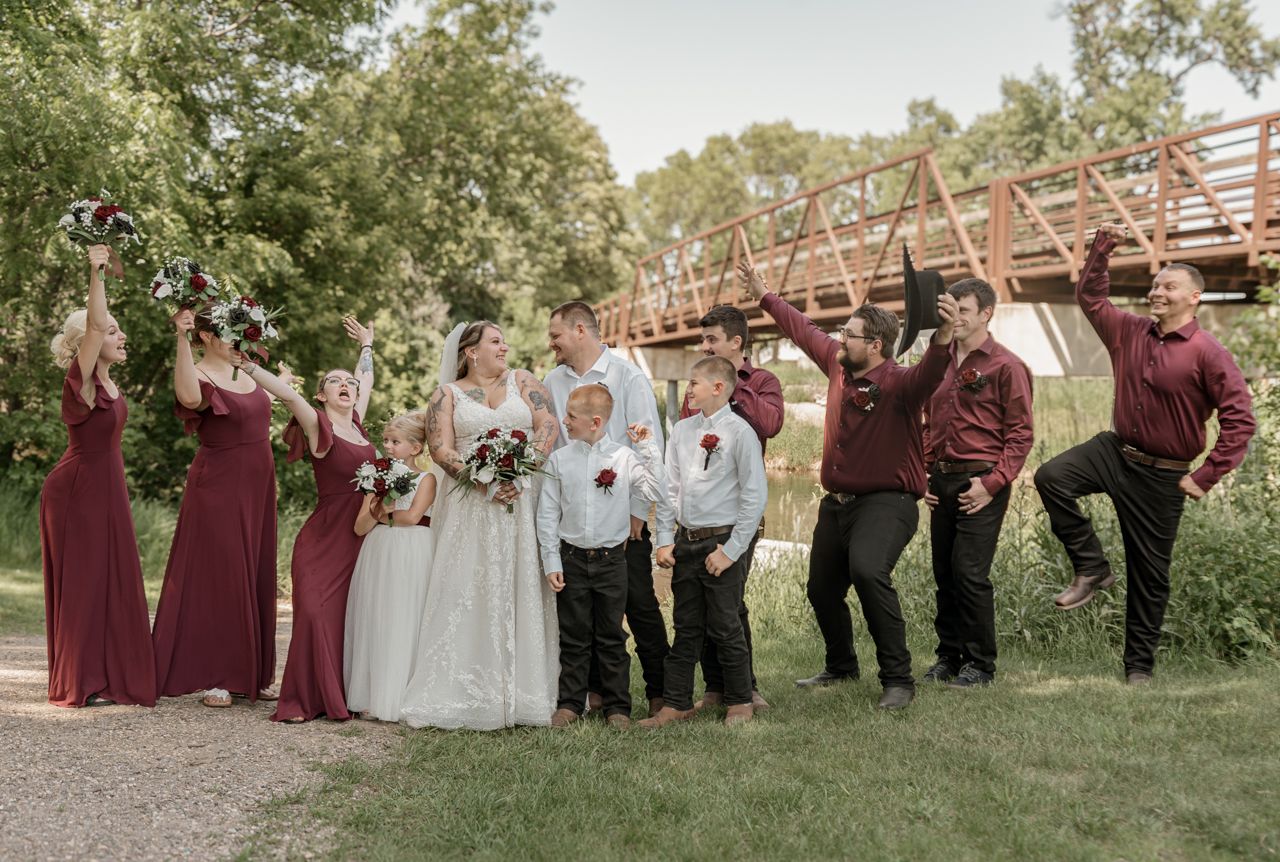 Wedding party poses on grassy hill near rustic wooden bridge in burgundy and white attire.
