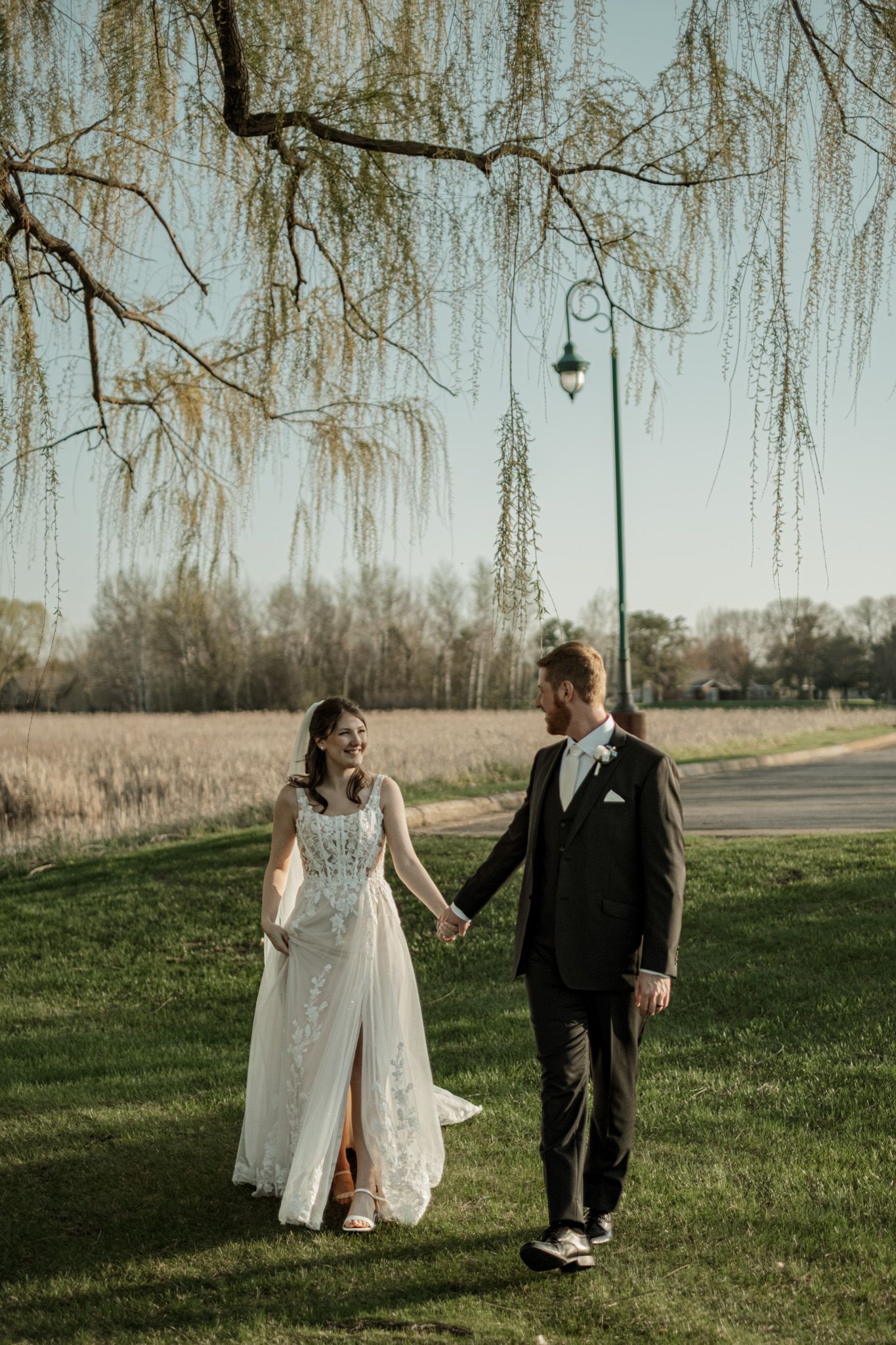 Newlywed couple walking hand in hand under willow trees in a romantic outdoor setting.
