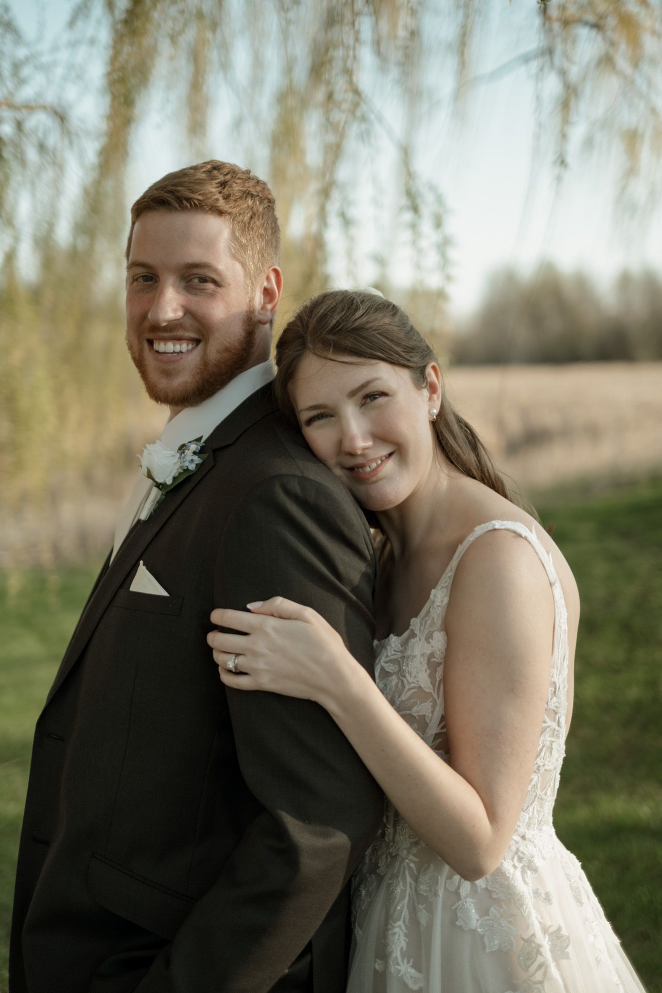 A newlywed couple poses together under willow trees in a romantic outdoor wedding portrait.