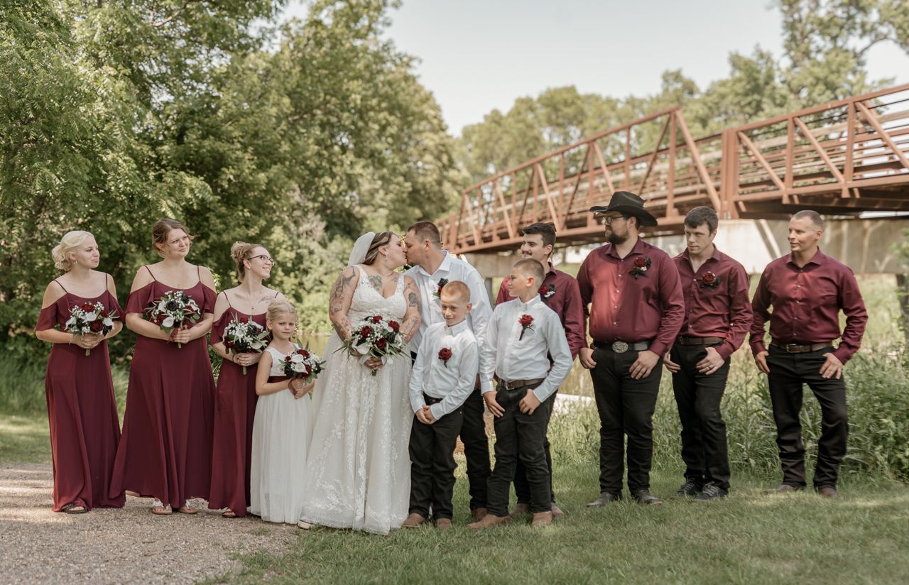 Wedding party in burgundy attire poses outdoors in front of rustic wooden bridge during fall ceremony.