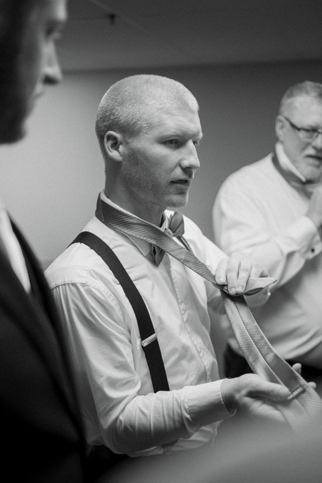 Black and white photo of someone adjusting their tie while wearing suspenders with others in a formal setting.