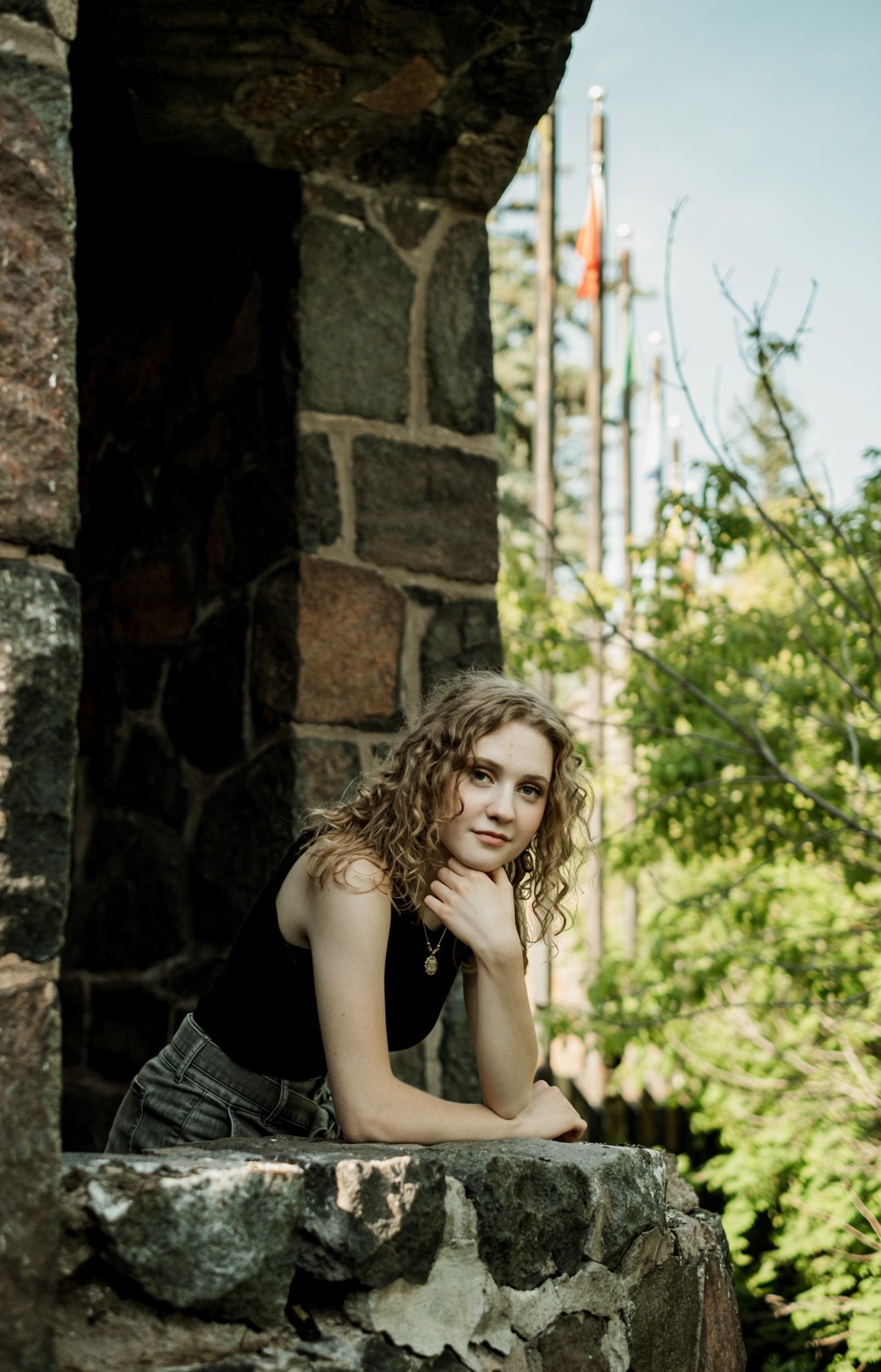 Figure seated on a stone ledge against a rustic stone wall with greenery in the background.