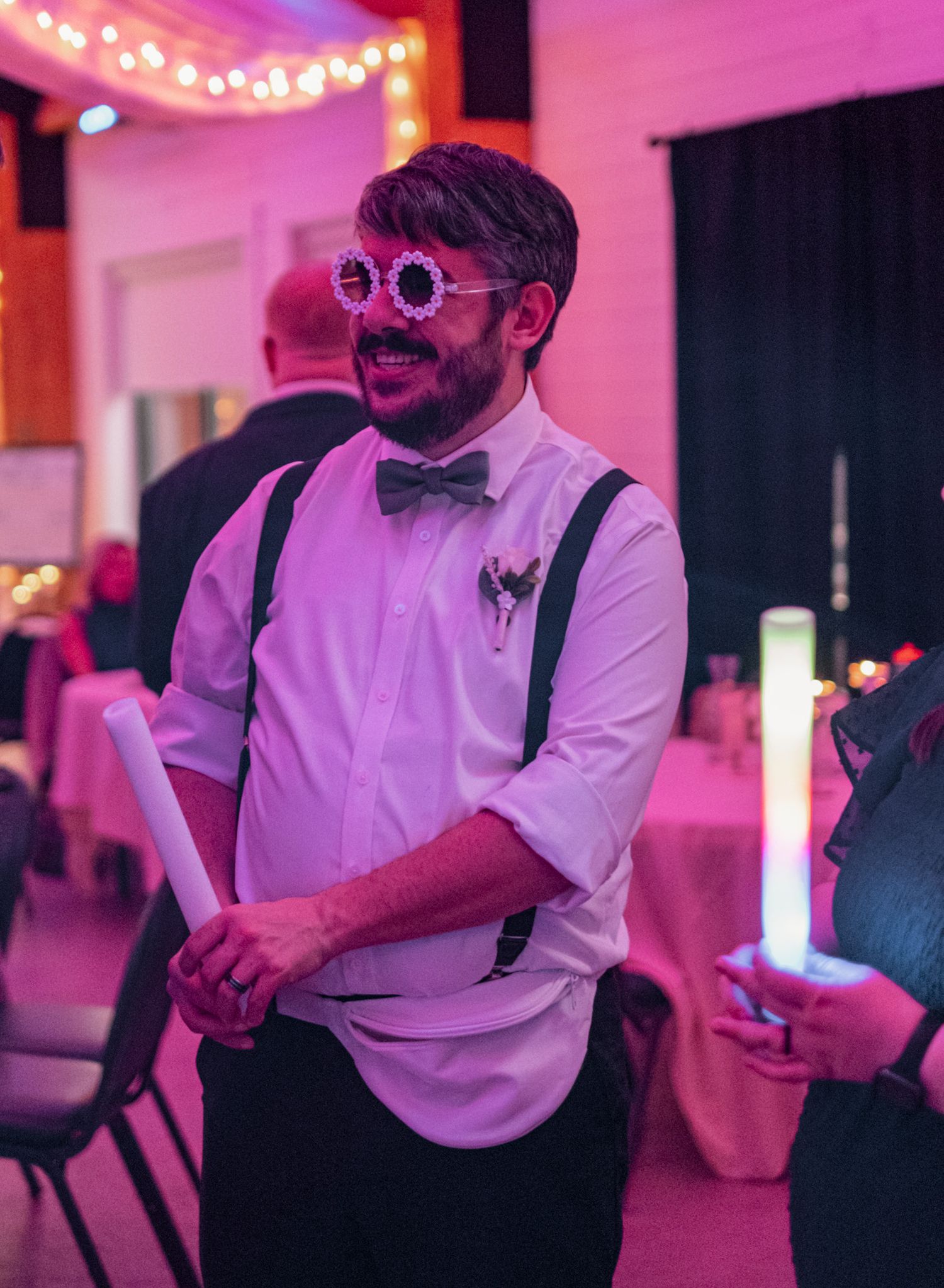 Person in suspenders and bow tie wearing round sunglasses smiles at party with pink lighting and string lights.