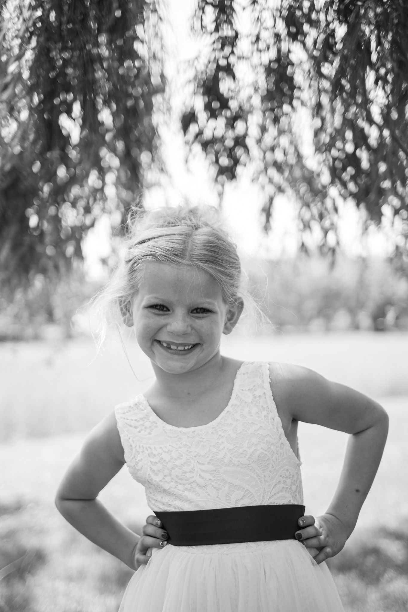 Black and white portrait of a young child in a white dress with black sash standing under tree branches.