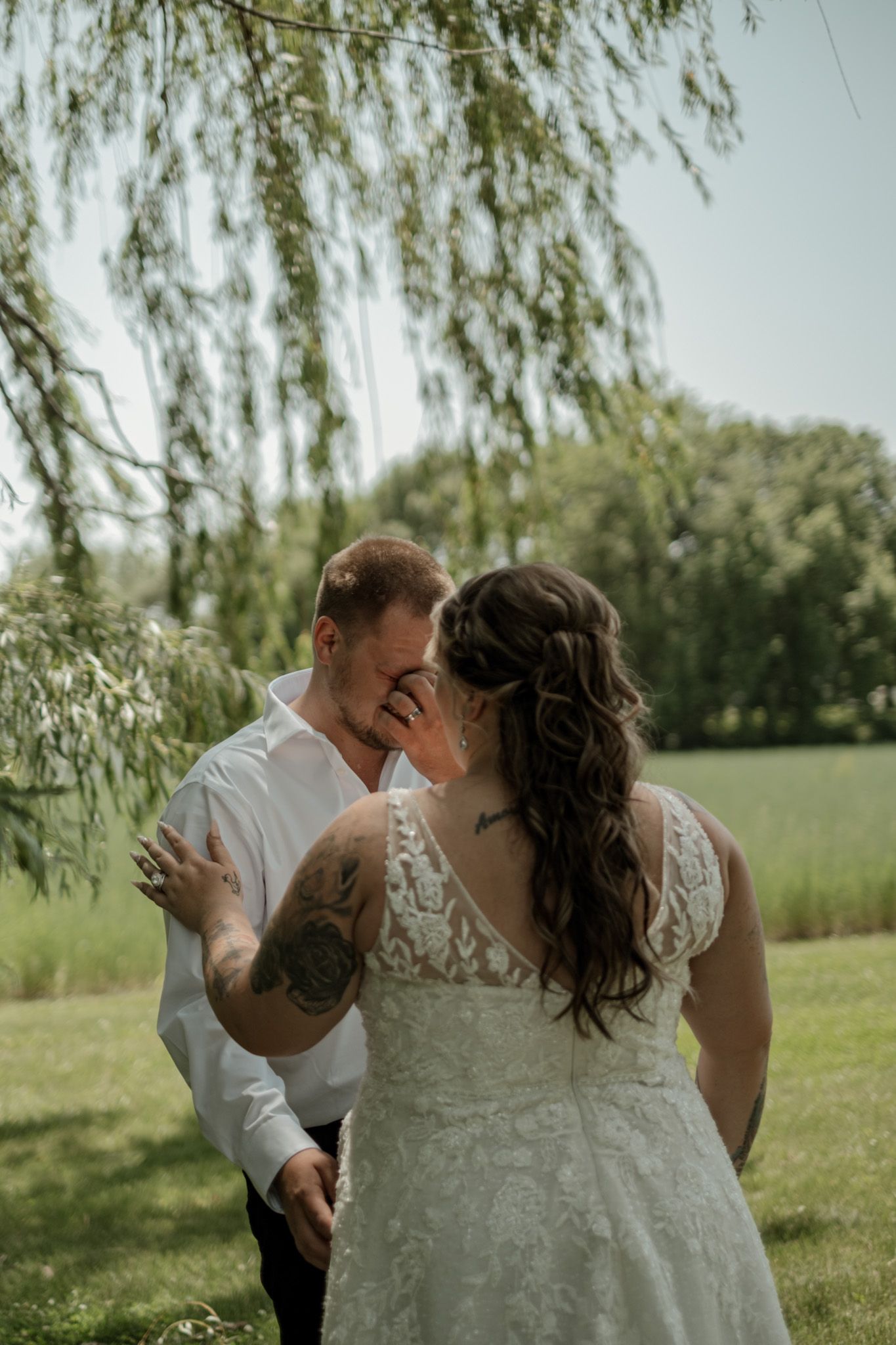 A romantic wedding moment under a weeping willow tree shows a couple sharing an intimate embrace outdoors.