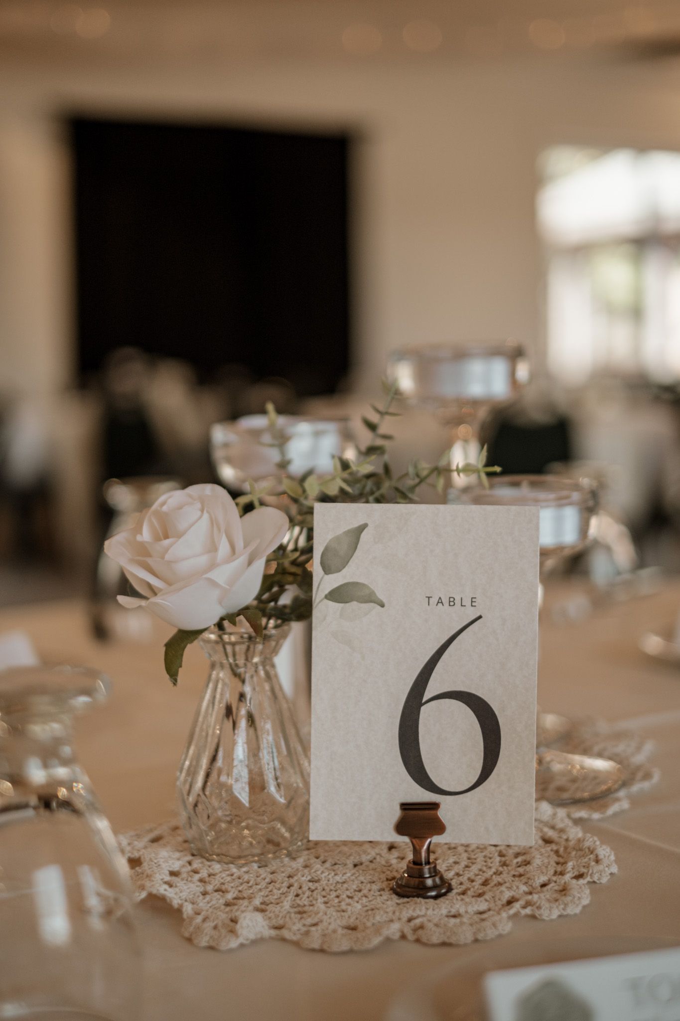 Elegant wedding table number six displayed on a doily with soft pink roses and greenery in a glass vase centerpiece.