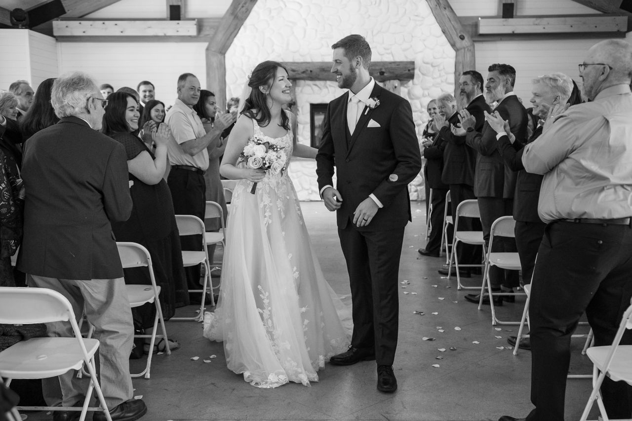 A newlywed couple walks down the aisle while guests clap and take photos.