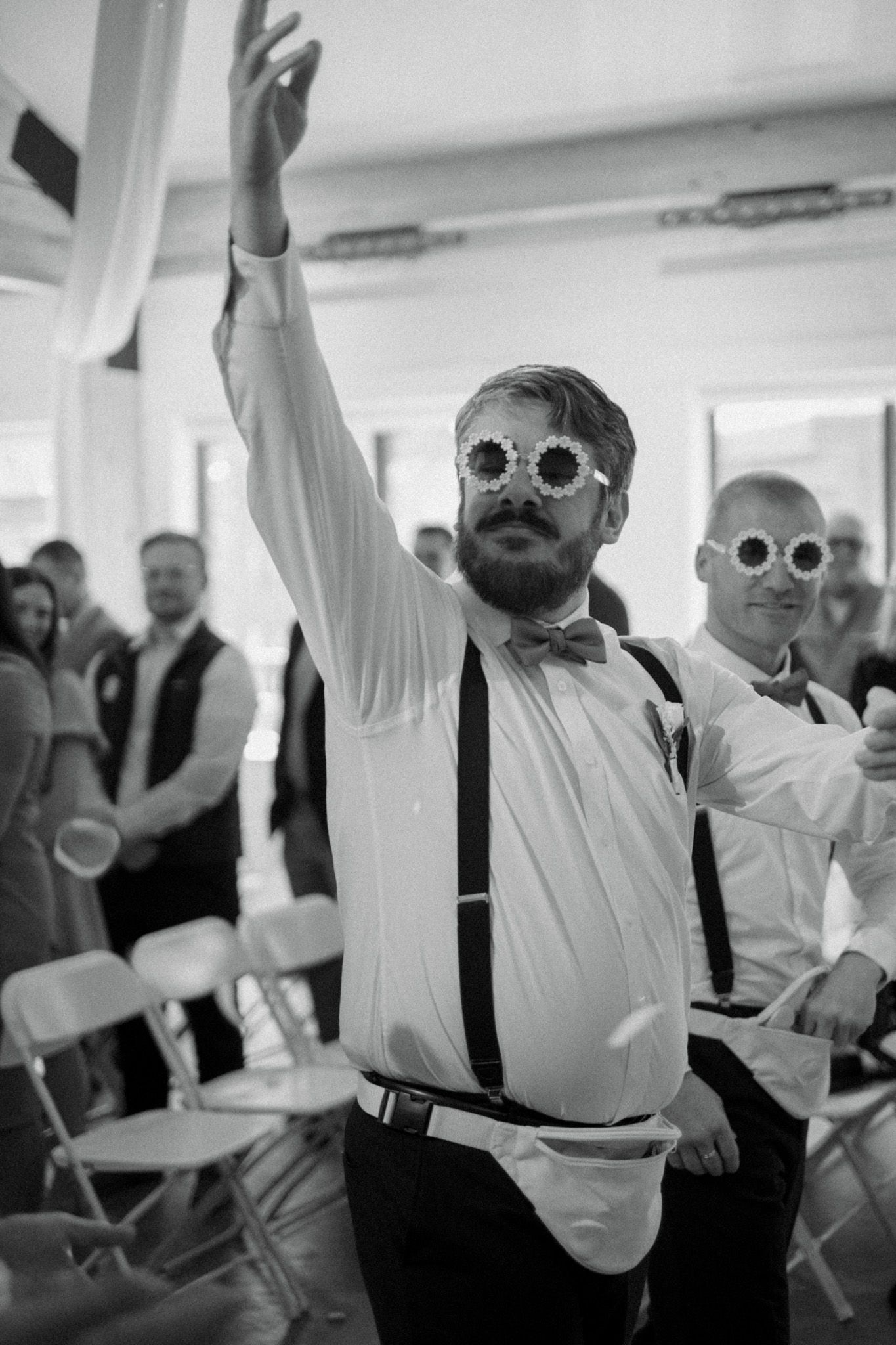 Wedding guest in suspenders and sunglasses dancing with raised arm at reception.
