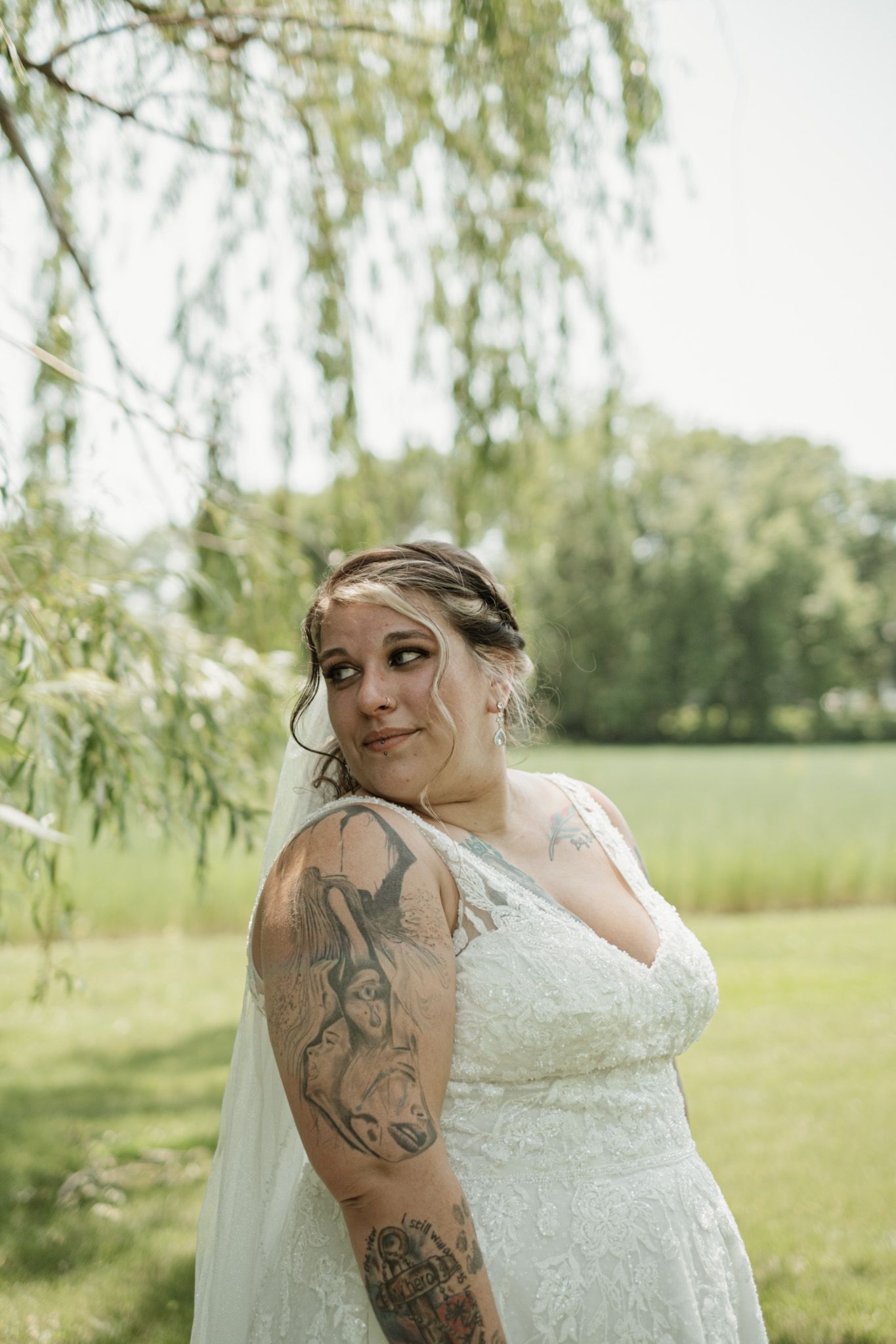 A bride in a lace wedding dress with visible tattoos poses beneath a willow tree in an outdoor garden setting.