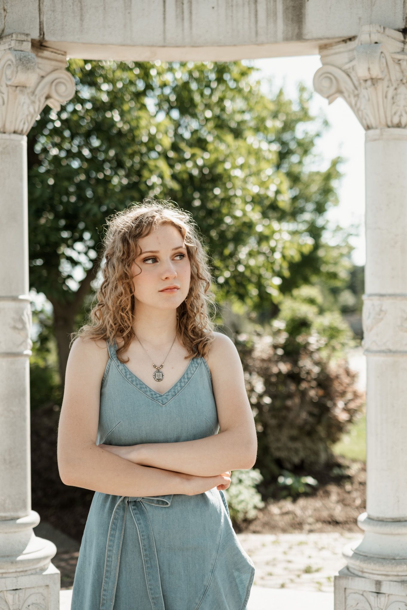A figure in a blue sundress stands between ornate white pillars on a sunny summer day.