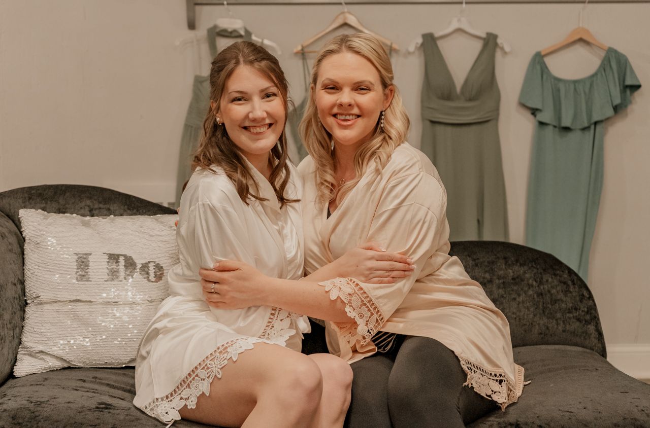 Two women in robes sit together on a couch during wedding preparations with bridesmaid dresses hanging behind them.