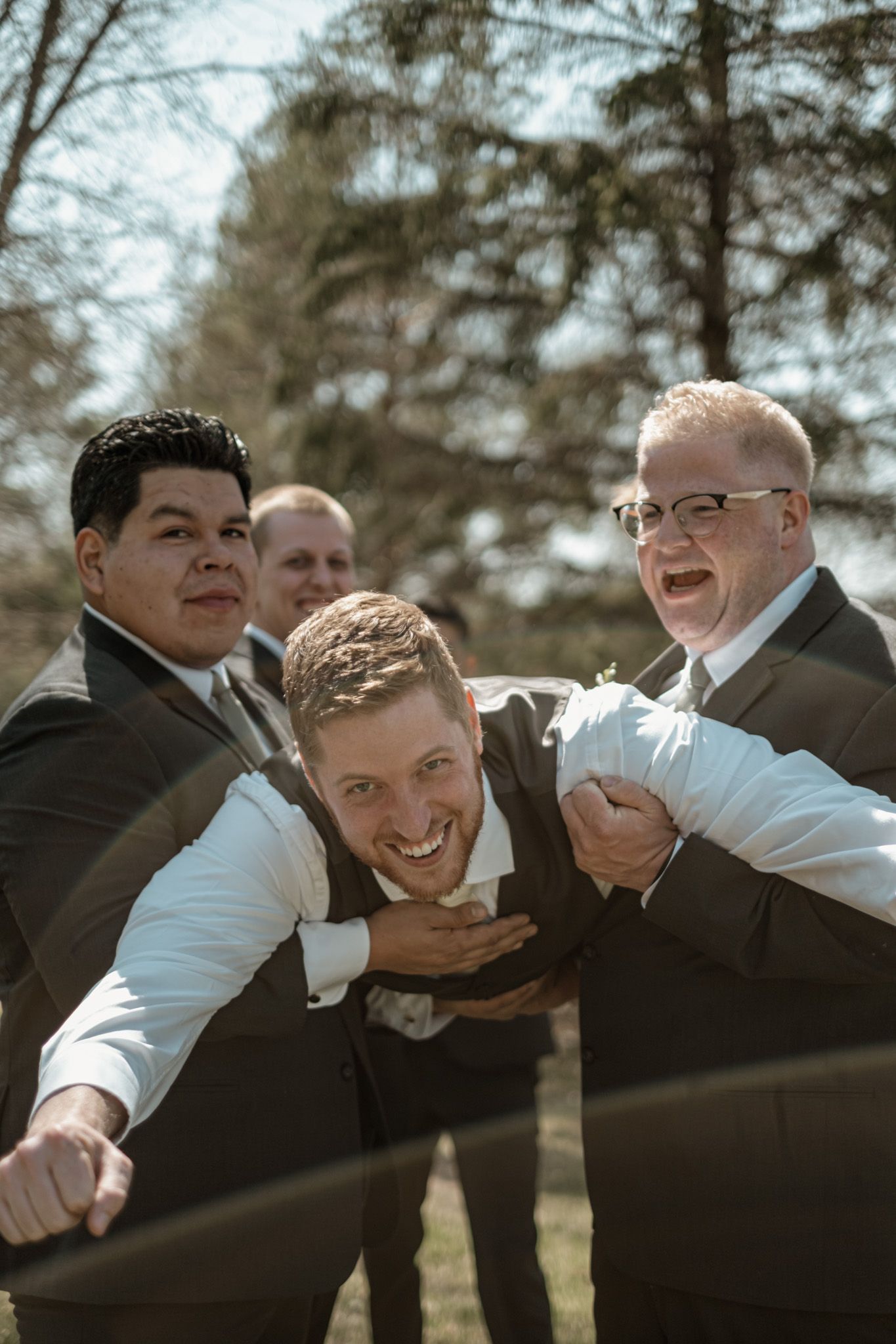 Three groomsmen in suits playfully posing and laughing together outdoors during a wedding photoshoot.