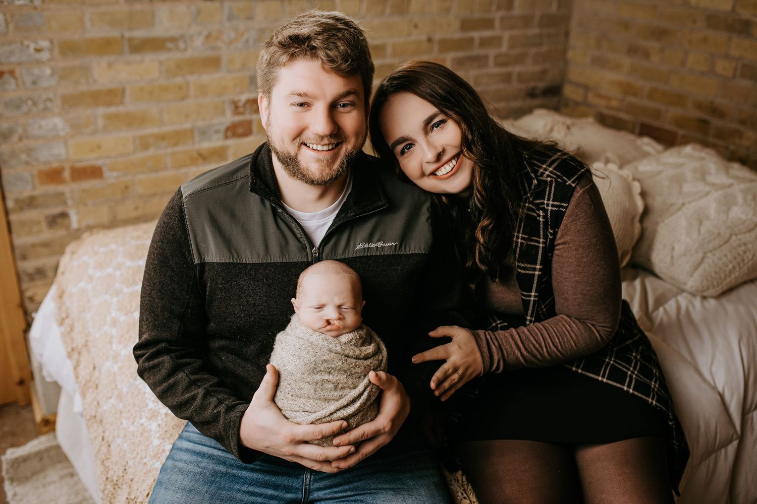 A couple sits together on a couch holding their newborn baby in a room with exposed brick walls.