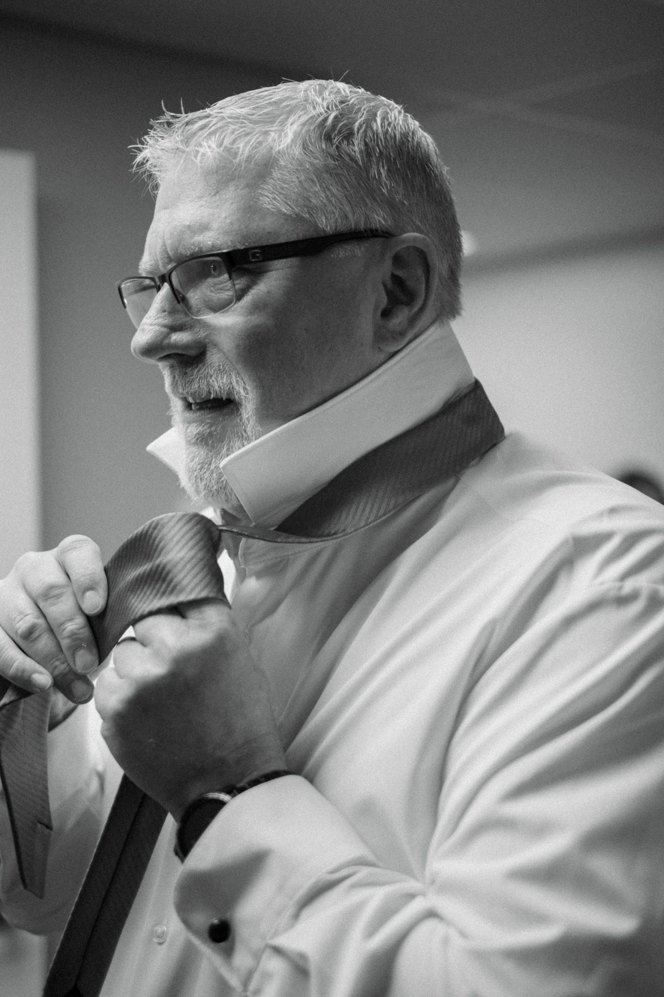 Black and white portrait of a gentleman in glasses adjusting his tie while wearing a white dress shirt.