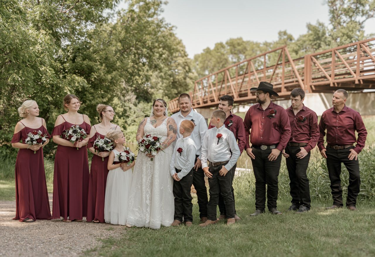 A wedding party in burgundy and white attire poses outdoors in front of a rustic wooden bridge on a sunny day.