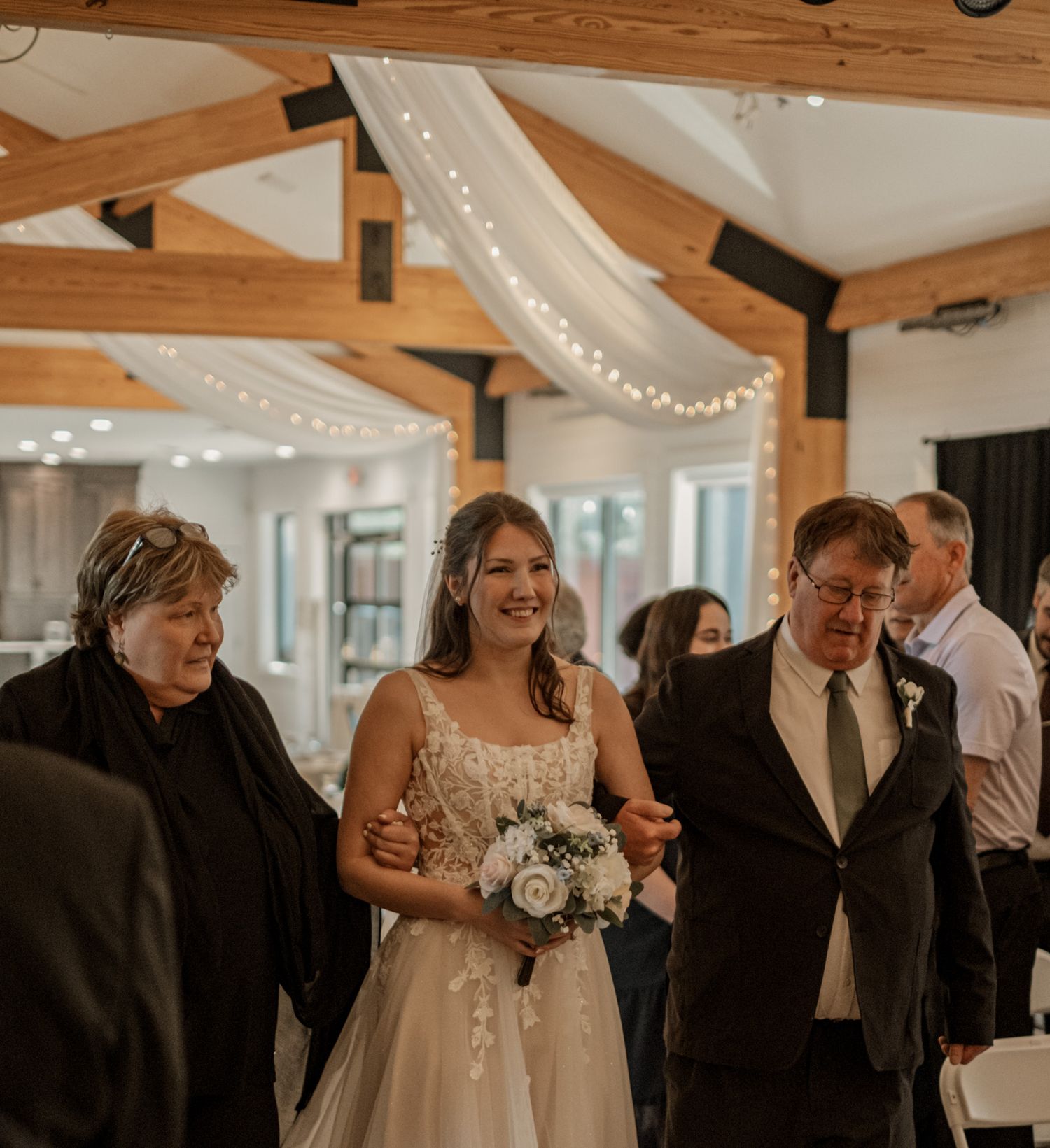 A wedding ceremony processional in a venue with wooden beam ceilings decorated with string lights.