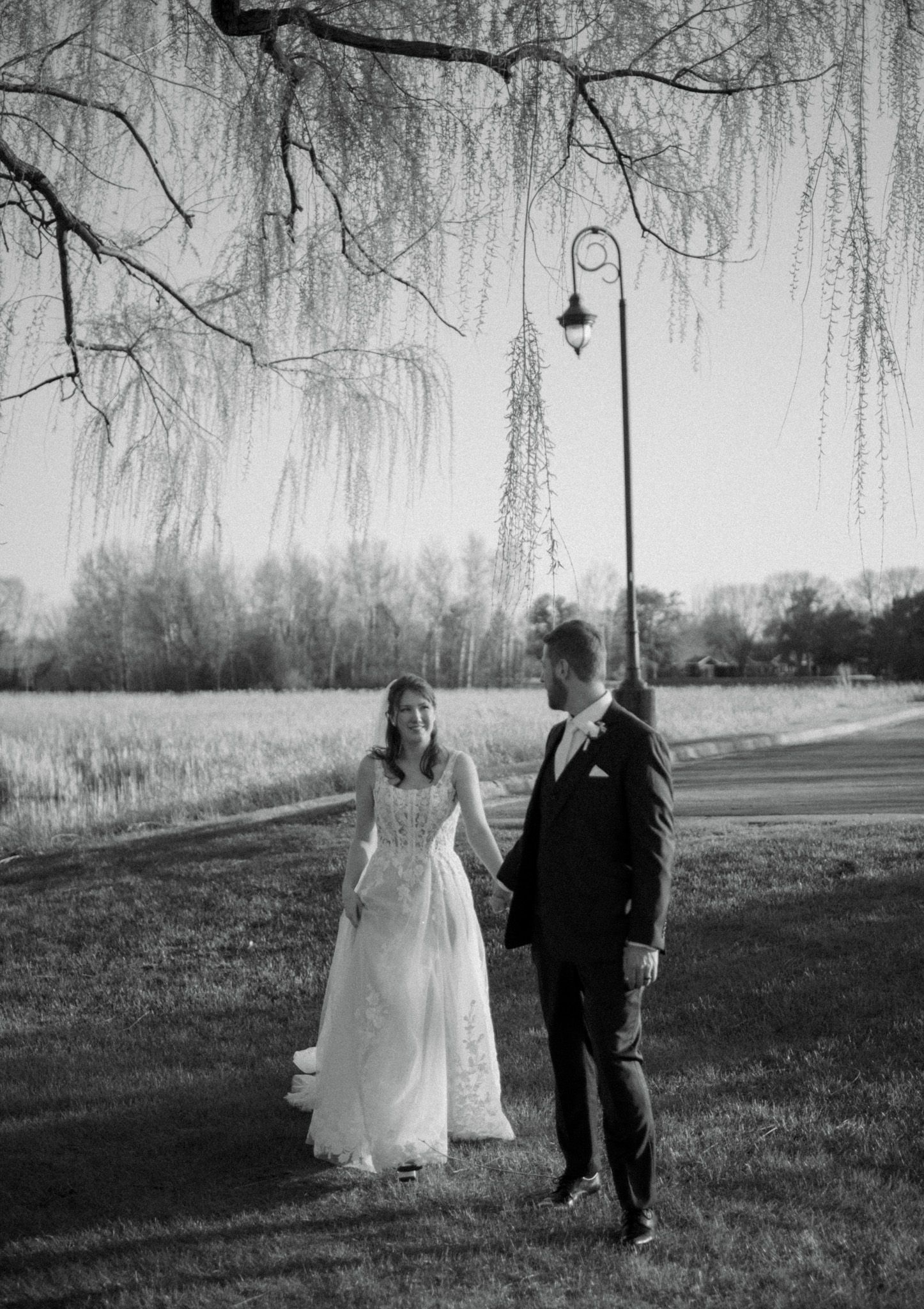 Black and white wedding photo of a couple standing under weeping willow trees near a vintage lamppost.