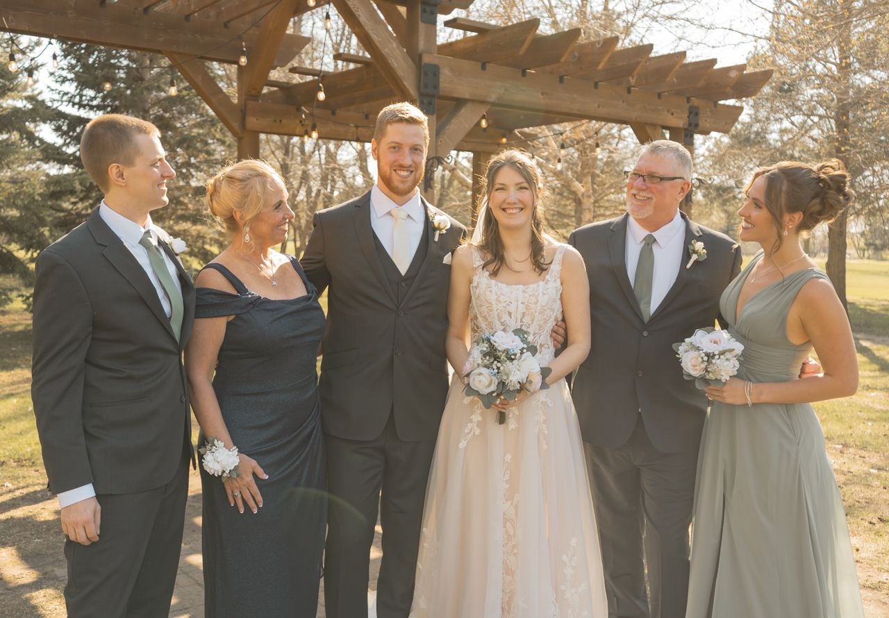 Wedding party poses outdoors under wooden pergola on sunny day.