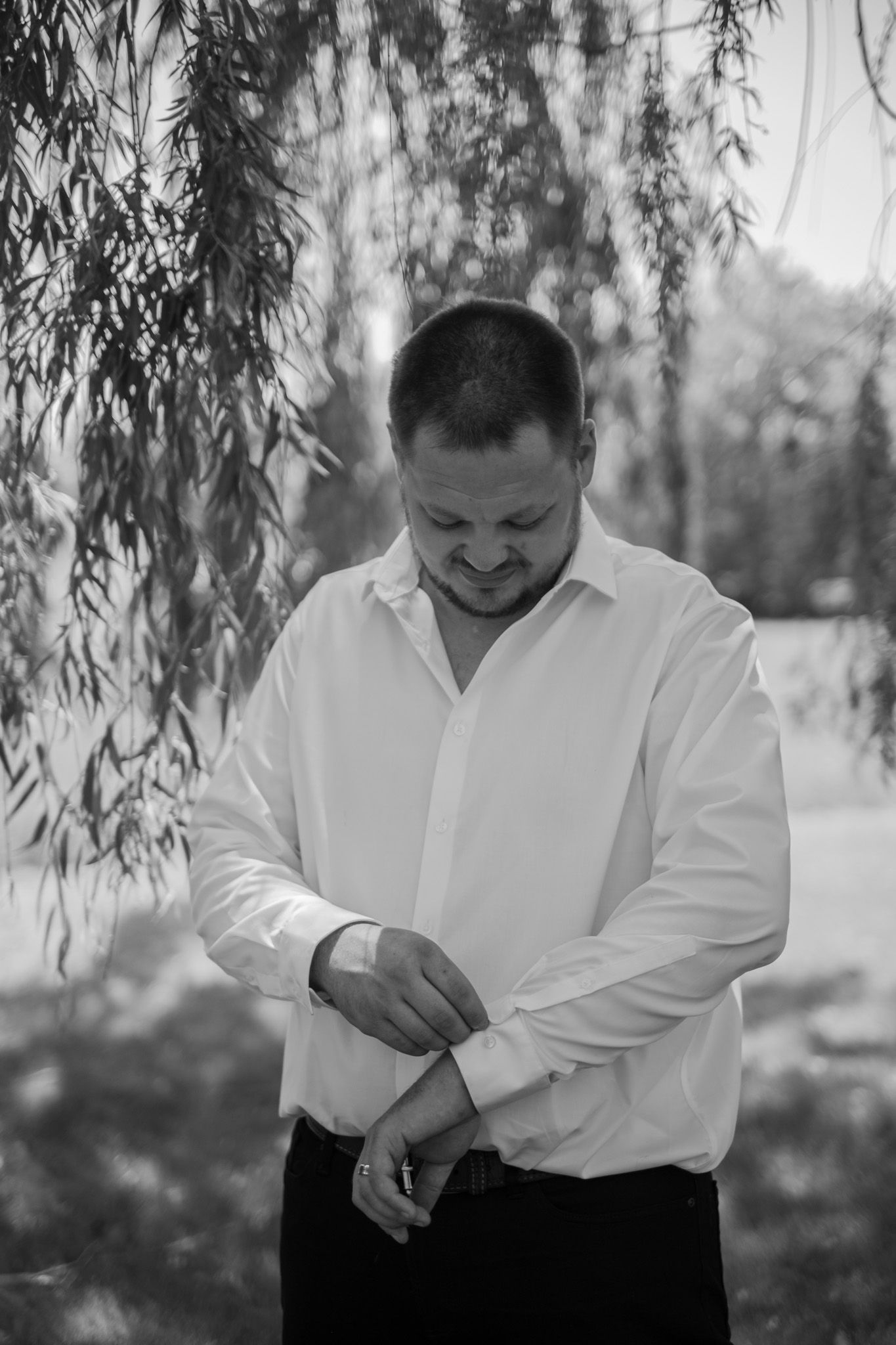 Black and white portrait of someone in a white dress shirt adjusting their sleeve under a willow tree.