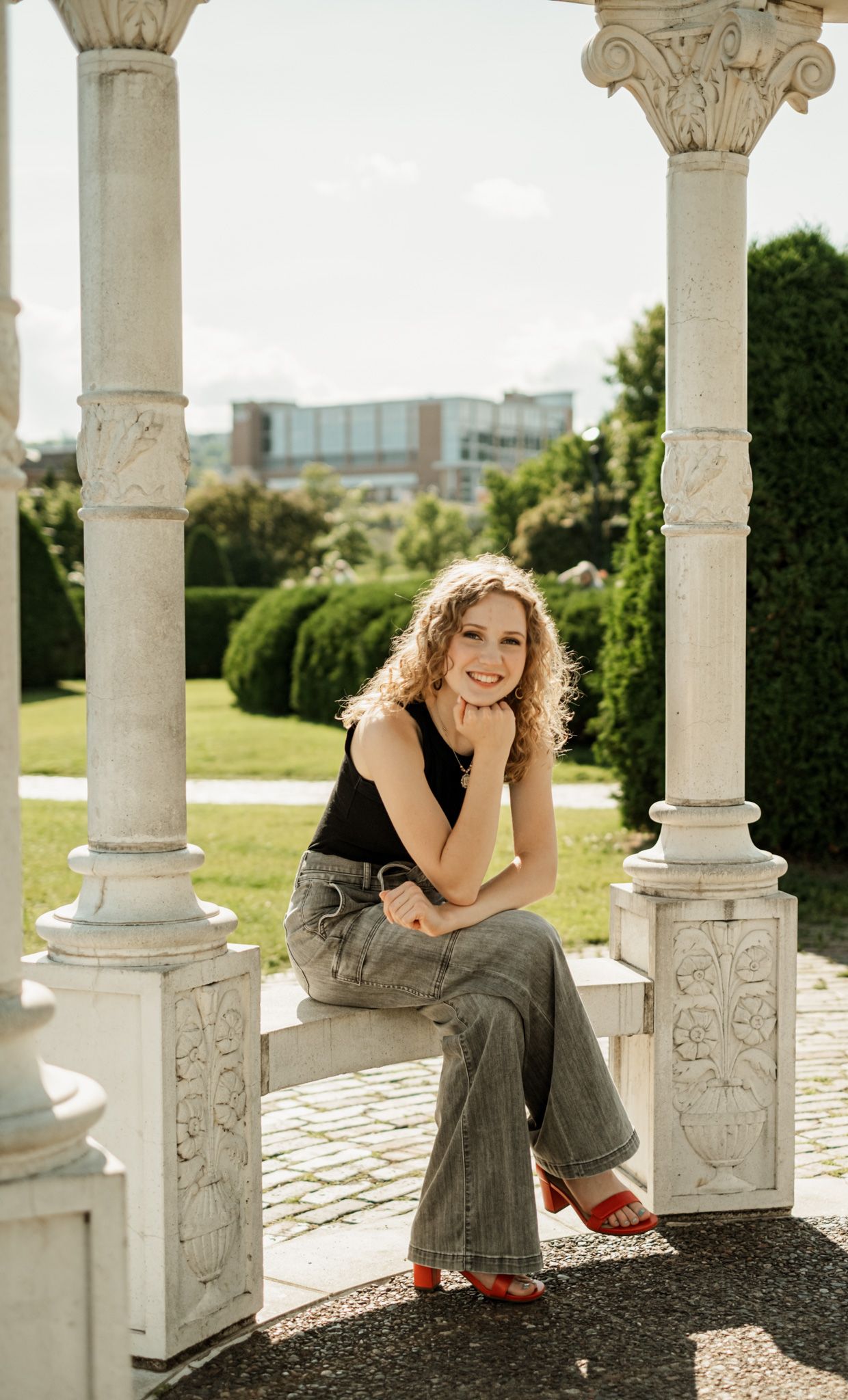 A woman sits casually between ornate white stone columns in an outdoor garden setting wearing casual attire.