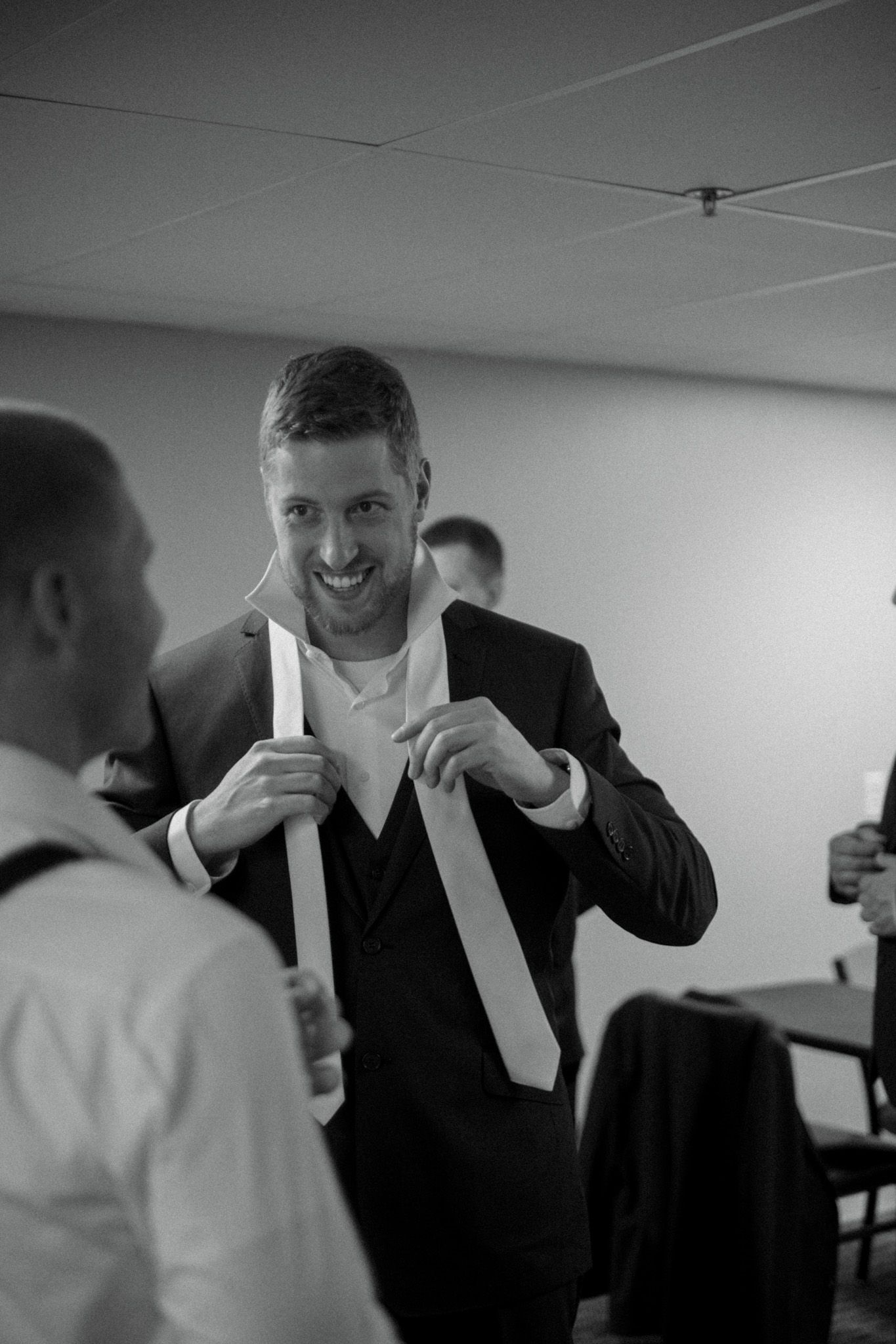 A groomsman adjusts his white tie while getting ready in a indoor hallway at a wedding.