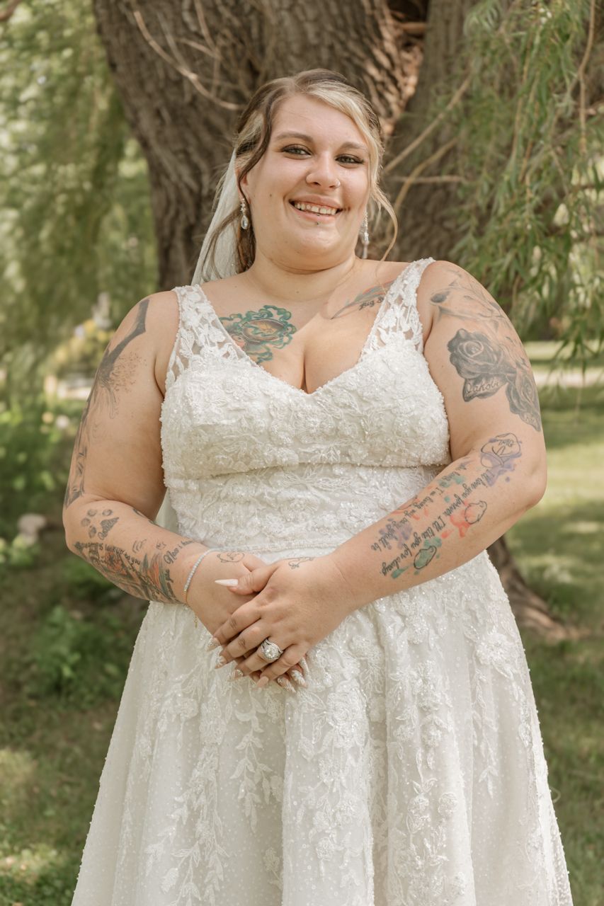 A radiant bride in a lace white wedding dress with visible tattoos poses against a tree in a garden setting.