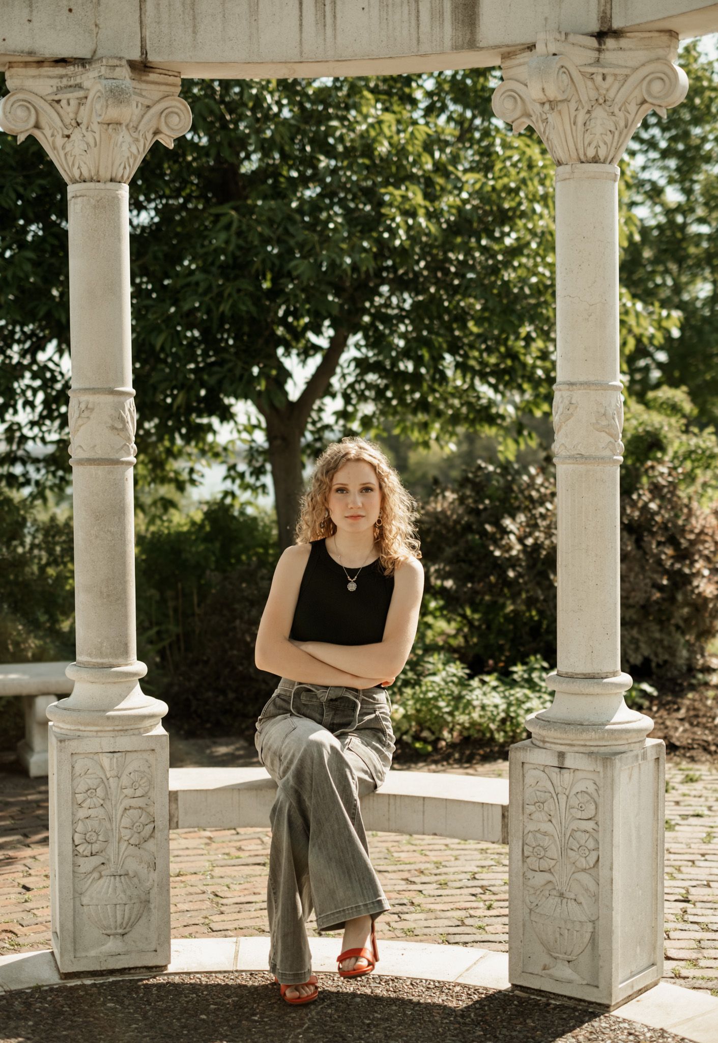 Stylish woman poses in casual attire against ornate stone columns and garden backdrop in golden sunlight.