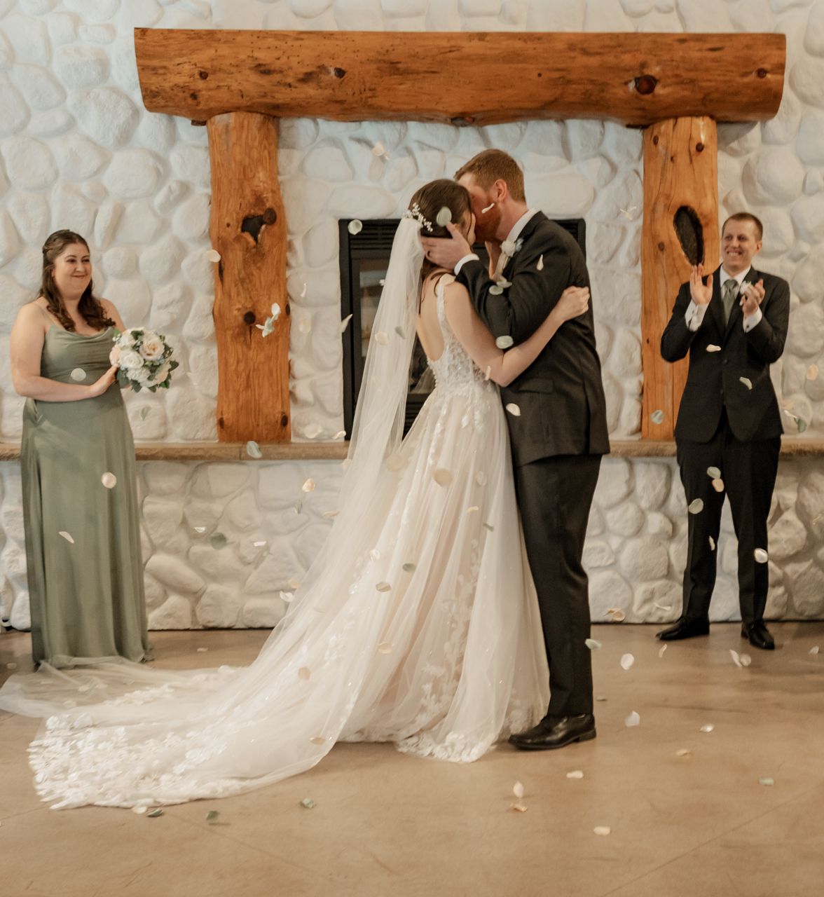 Wedding ceremony under rustic wooden arch with stone backdrop and wedding party looking on.
