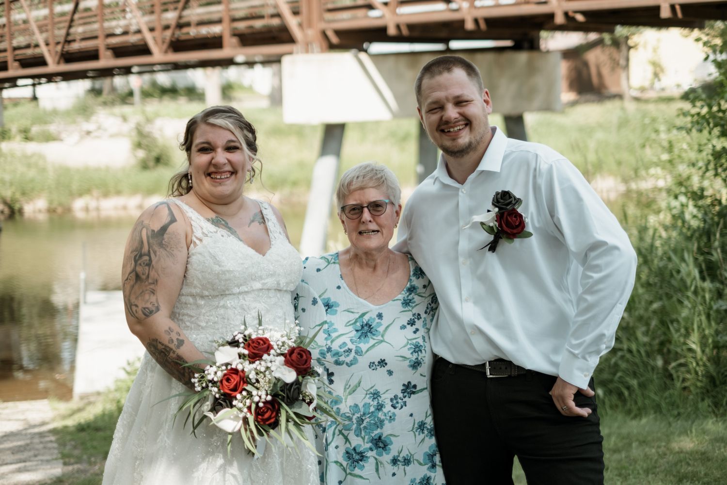 Wedding party posing together under a metal bridge on a sunny summer day near a river.