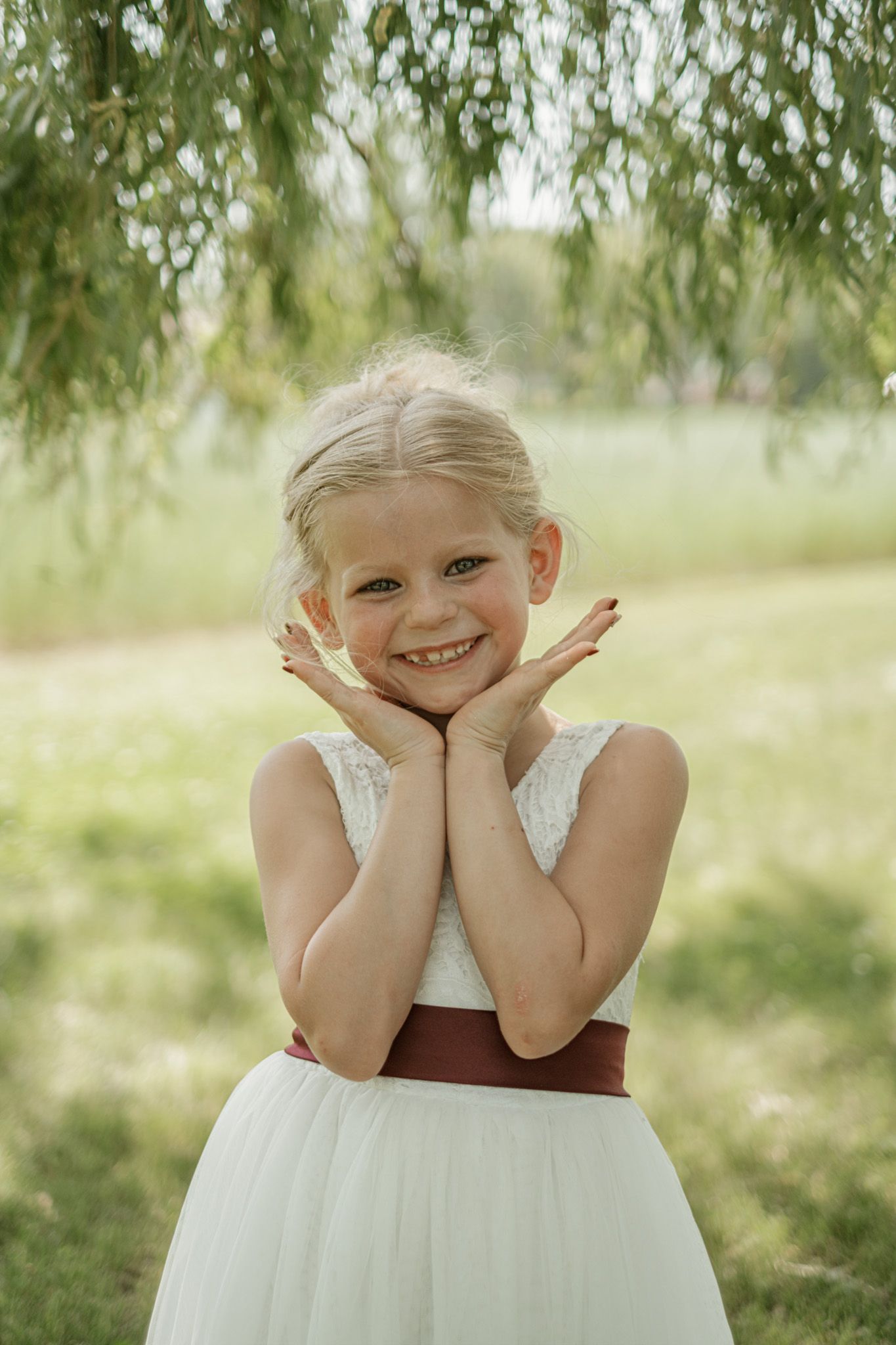Child wearing white floral dress with brown sash poses outdoors under willow tree branches in natural setting.