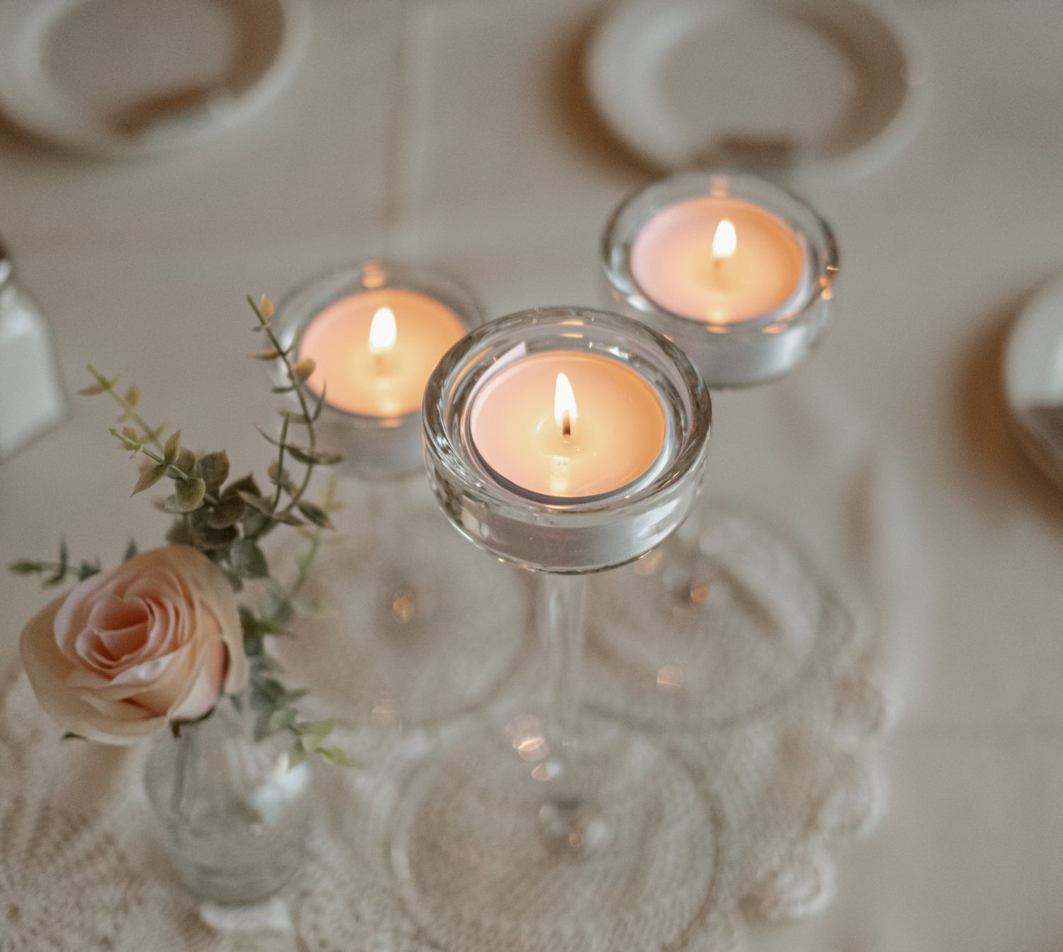 Lit tealight candles in glass holders with a delicate pink rose centerpiece create an elegant table setting.