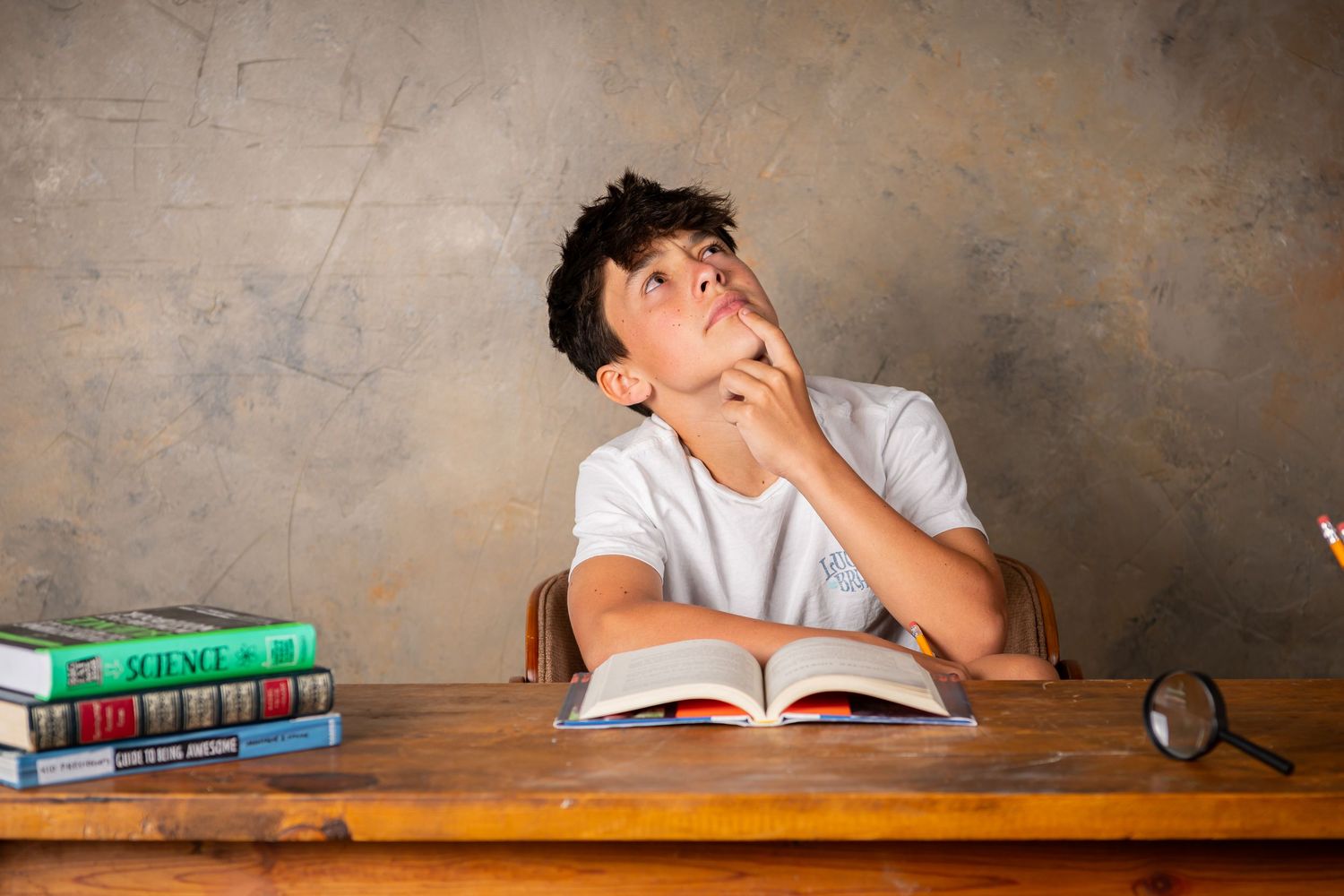 Student appears exhausted and stressed while studying at wooden desk with stack of textbooks beside them in child photography session Omaha.