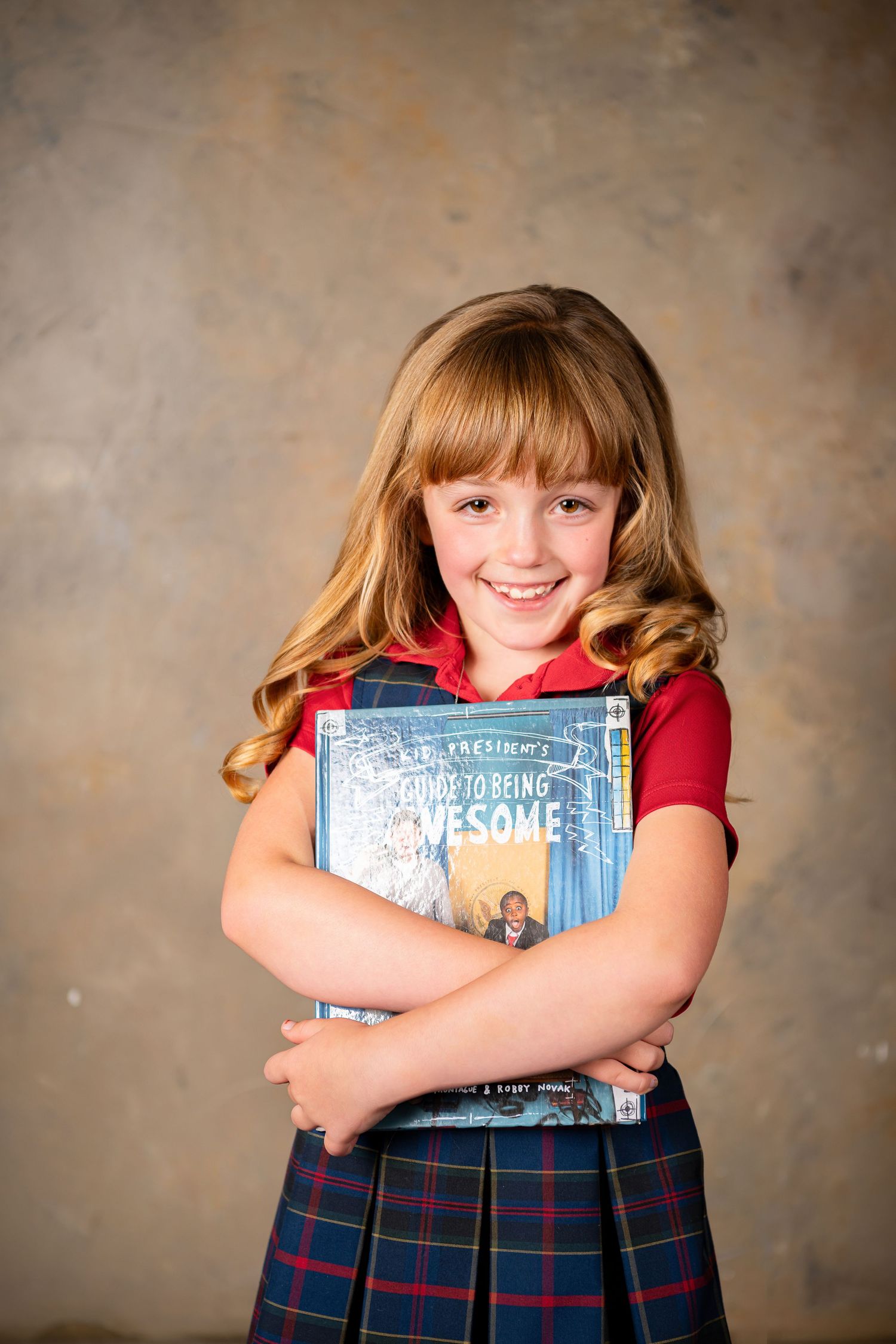 Student in plaid school uniform hugs book while smiling against textured brown background in child photography session Omaha, Nebraska.