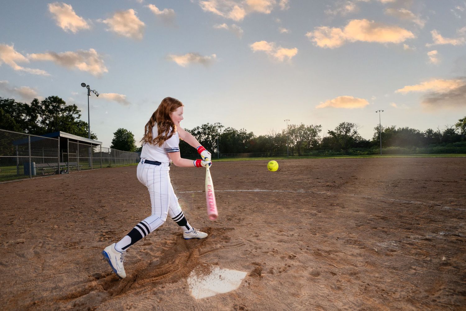 Softball player in white uniform swings bat on dirt field during sunset with cloudy sky in background taken by Omaha sports photographer.
