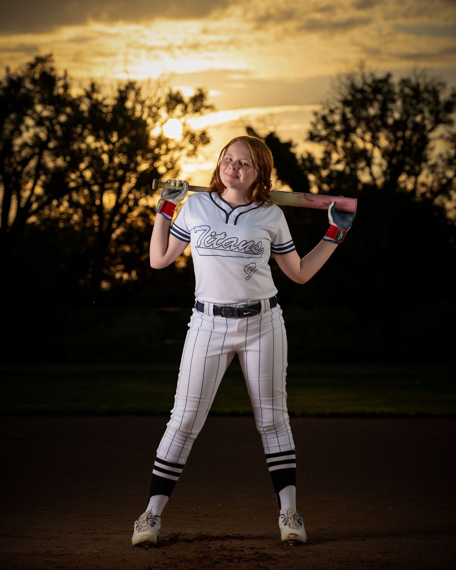 Baseball player in pinstriped uniform poses with bat at sunset on field with dramatic golden sky in background in Iowa sports photo session.